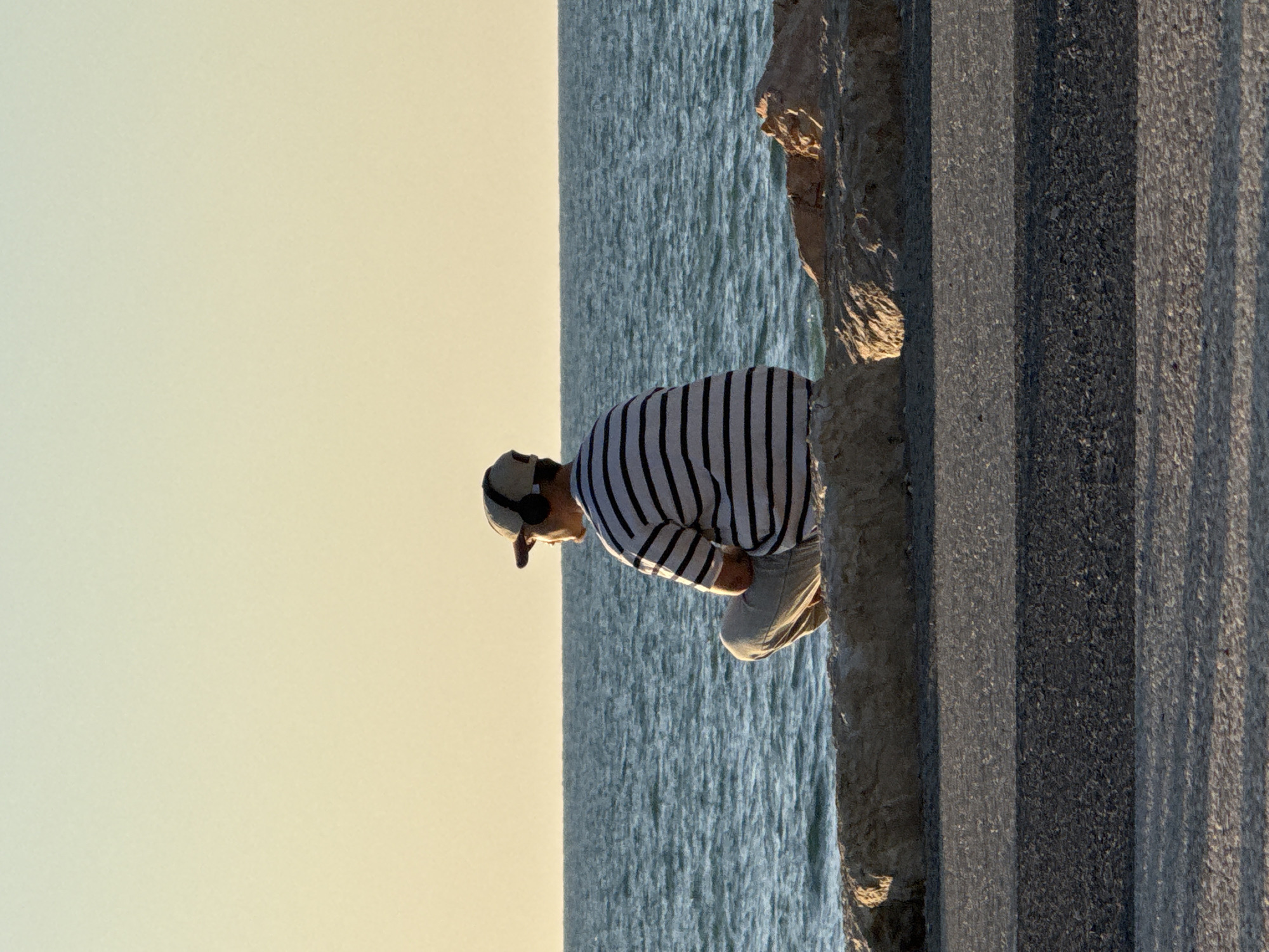 A lone individual is captured by the sea during what appears to be the golden hour, suggesting either sunrise or sunset. The person, seen in profile, is a male figure wearing a white and black striped long-sleeved shirt, light-colored trousers, a light-colored cap, and over-ear headphones. He is in a crouching or squatting position on a rugged, concrete or rocky ledge, gazing downwards towards the water.
The background consists of a serene expanse of calm, blue sea, showing subtle ripples on its surface. Above the water, a clear sky transitions from a pale yellow-cream hue at the horizon to a slightly warmer tone higher up. To the right of the person, a dark grey, textured paved area or wall with distinct vertical lines and shadows implies a constructed promenade or coastal barrier. The warm, soft light of the low sun illuminates the person and the edges of the surrounding structures. The scene conveys a sense of quiet observation or contemplation by the water. There is no visible text in the image.