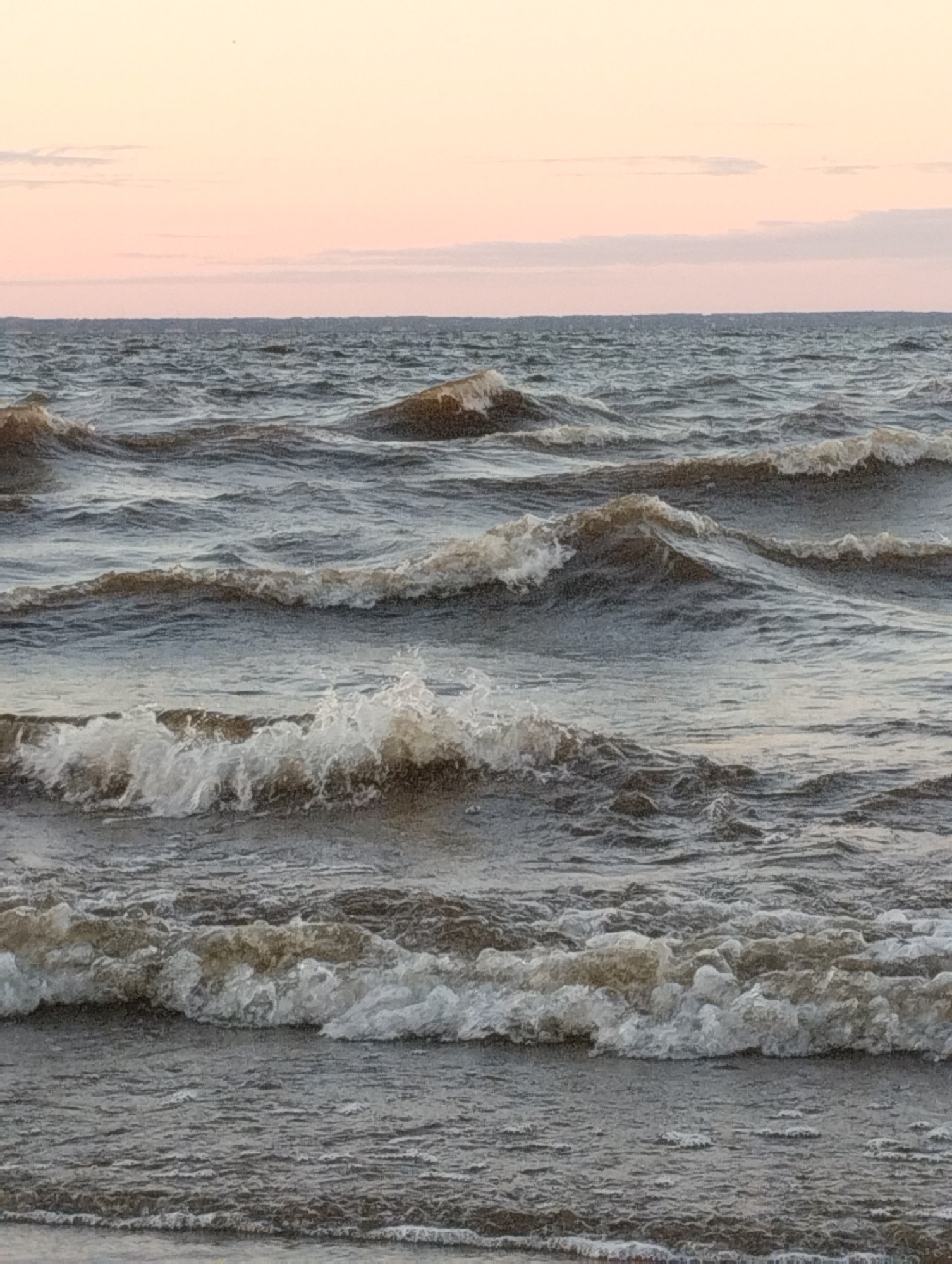A close-up, eye-level shot captures the dynamic motion of dark, choppy waves rolling towards the viewer under a pale pink and peach sky. The sky is smooth with a few subtle horizontal cloud bands. The water appears to be a large body, possibly a lake or sea, with white foam cresting on several waves. The composition is horizontal, with no land features visible except for a hint of a darker line on the horizon that could be distant land or a shoreline. The image is devoid of any human presence or man-made objects, focusing solely on the natural elements of water and sky.