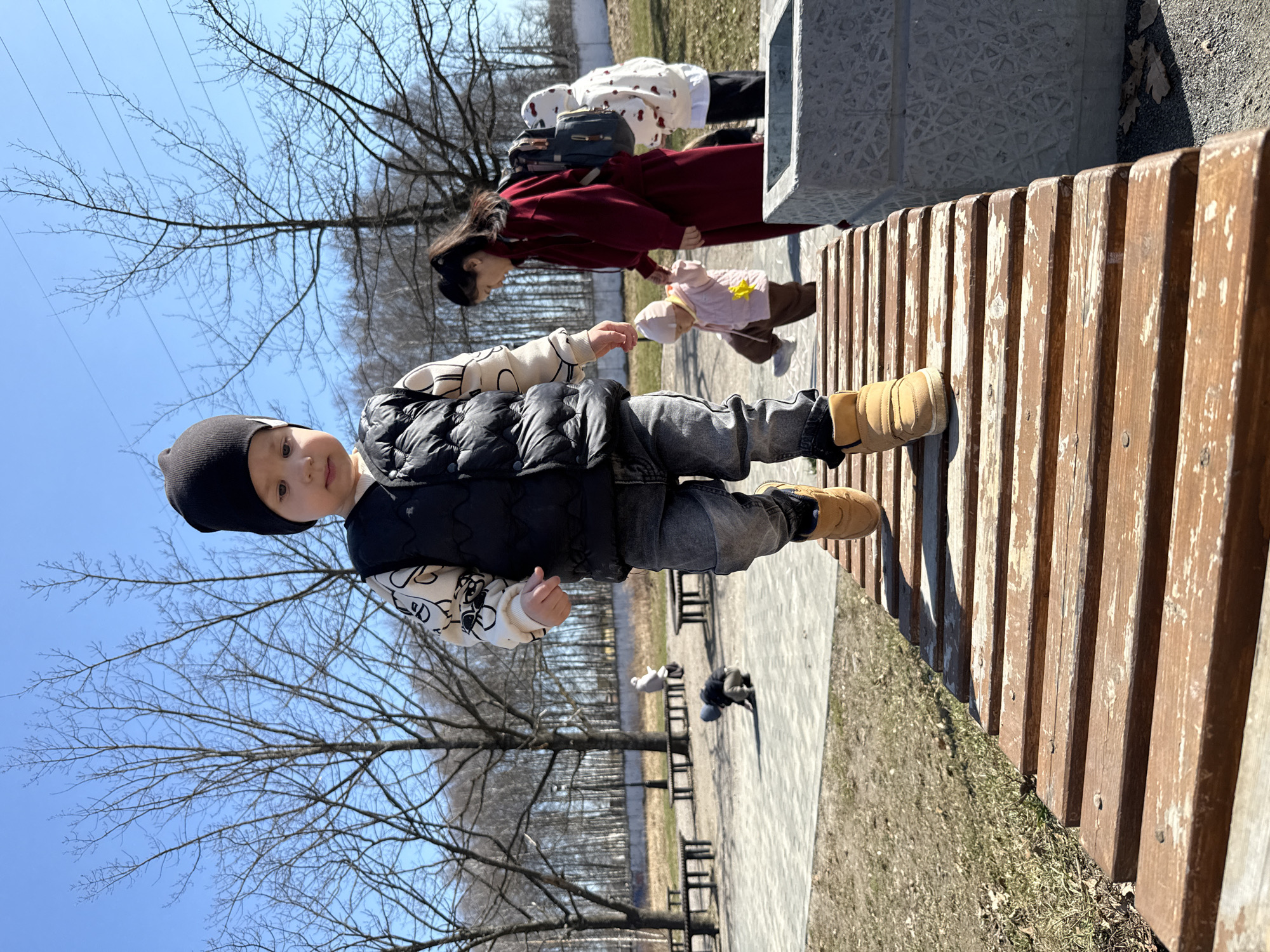 The image shows a child standing on a wooden bench in a park. The child is wearing a black beanie, a patterned white sweater, a black vest, gray pants, and tan boots. In the background, a woman in a red coat is seen holding a smaller child wearing a pink outfit. Other park benches are visible in the background, along with trees and a clear blue sky. Pigeons are on the ground. The location is Rusanovo, Russia.