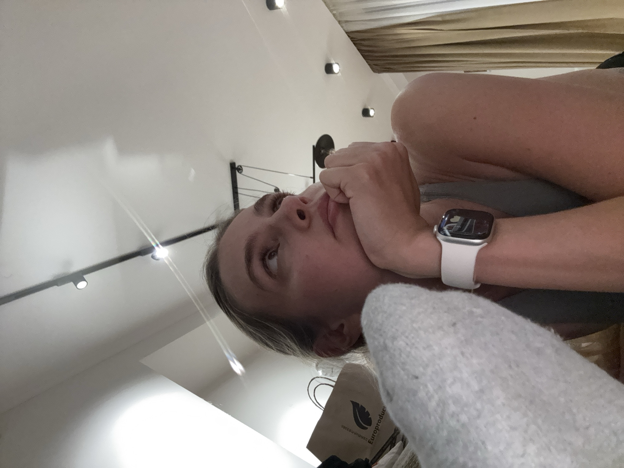 A woman is lying down, looking upwards with her hand near her face. She is wearing a smart watch on her wrist. A light gray, possibly knitted, object is in the foreground. A brown shopping bag with an indiscernible logo is partially visible. The ceiling has track lighting with multiple spotlights. The location is Unknown, Unknown.