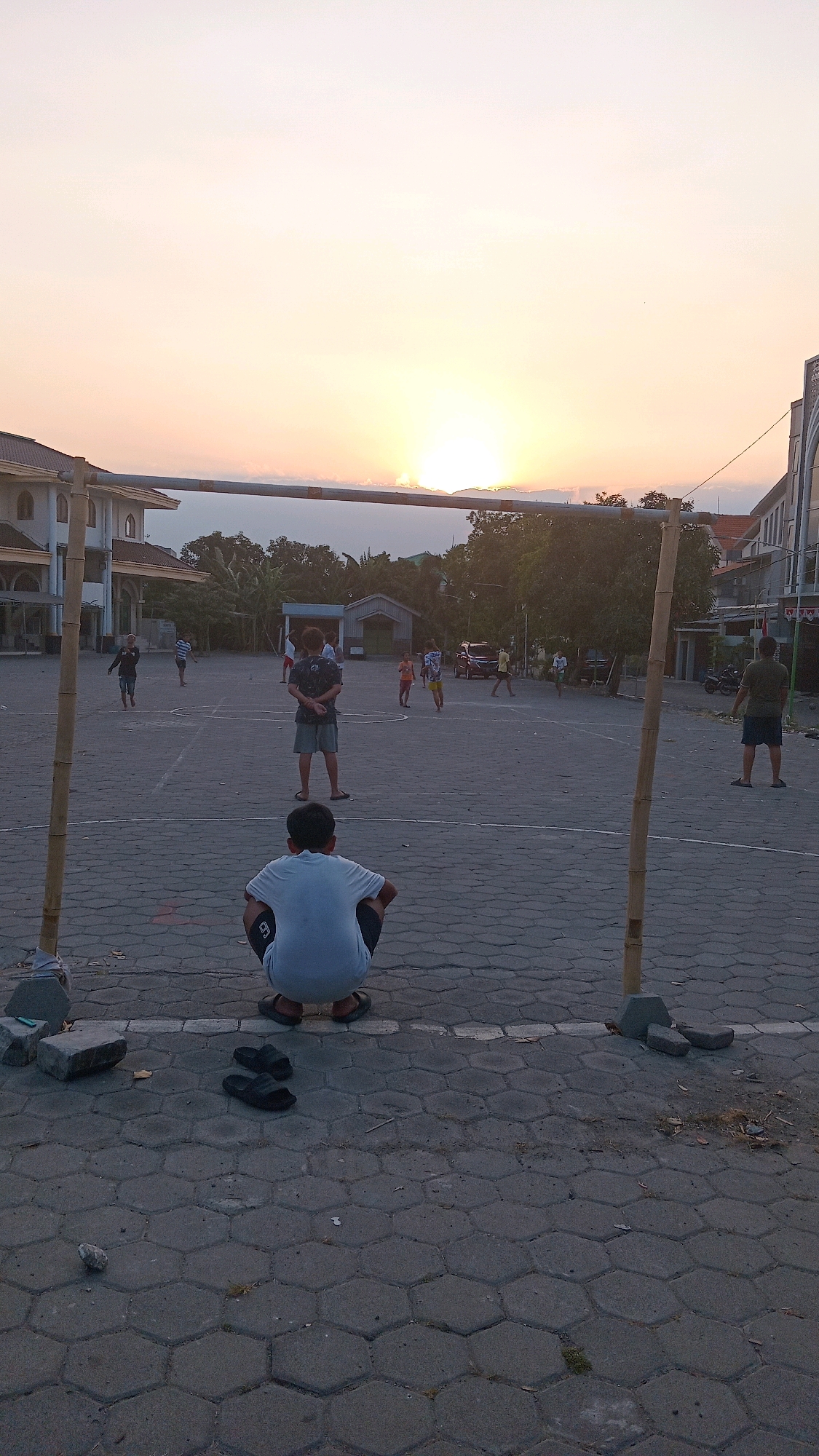 In Kembangan, Indonesia, at sunset, several young males are playing soccer on a paved court. The setting sun casts a warm glow across the sky, with its rays visible above the horizon. A makeshift soccer goal, constructed from bamboo poles and a horizontal bar, stands in the foreground. In front of the goal, a young boy wearing a white t-shirt and dark shorts crouches down, facing the game. Other players are scattered across the court, engaged in the match. The court is marked with white lines, and the surrounding area features buildings, trees, and a parked car. The ground is paved with hexagonal concrete blocks.