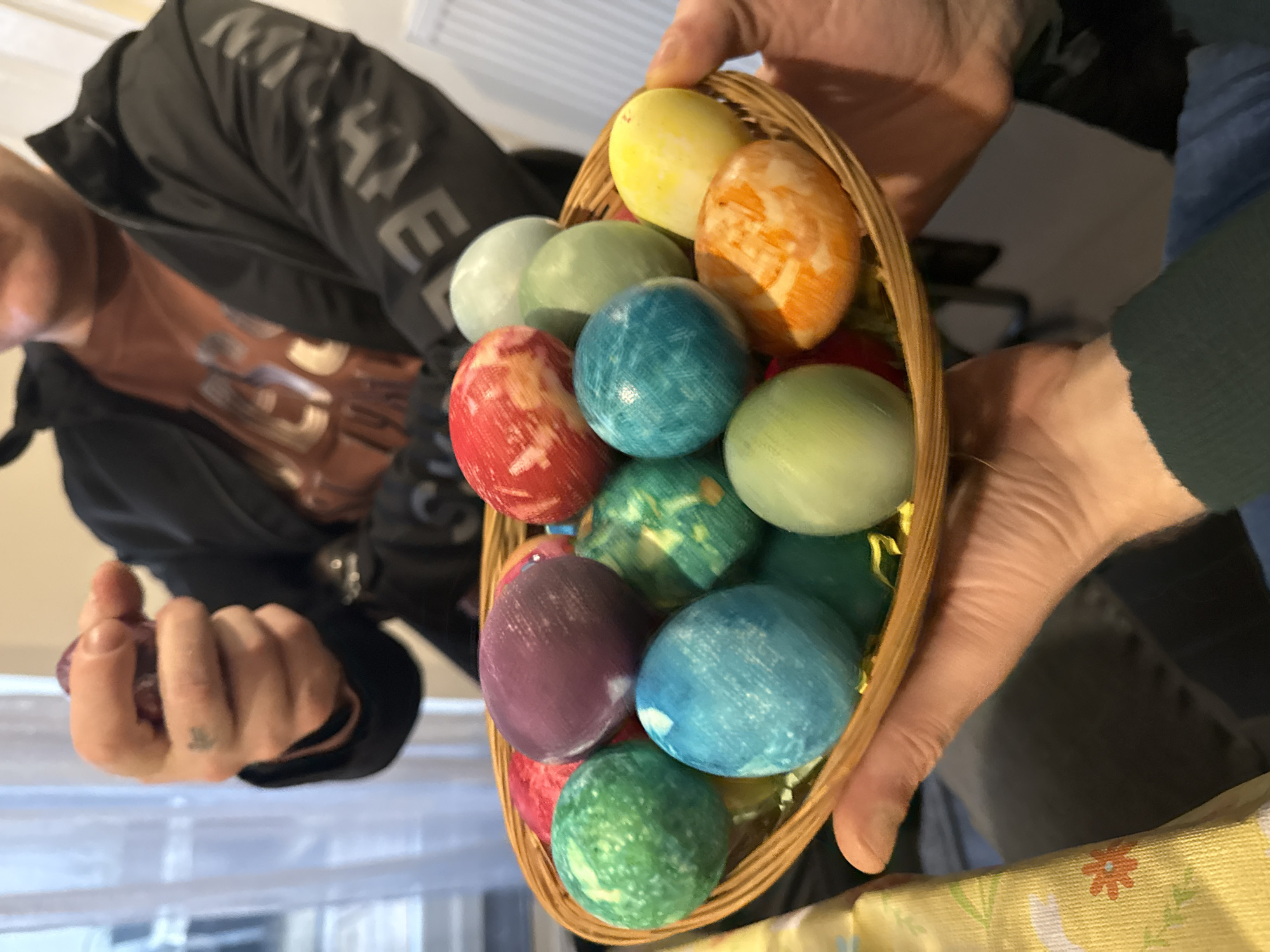 A basket of colorful Easter eggs is held in two hands. The eggs are dyed in various colors and patterns, including blue, green, yellow, orange, red, and purple. The basket is made of woven material. The hands holding the basket appear to belong to two different people, based on the sleeve colors. One person is wearing a black jacket and the other is wearing a blue shirt with a dark green cardigan. A portion of one person's face is visible. A third hand wearing black sleeve in holding an additional Easter egg. In the background, a window with sheer curtains is visible. The photo appears to be taken indoors, with a patterned tablecloth visible at the bottom of the frame. The location is not mentioned or able to be inferred from the image.