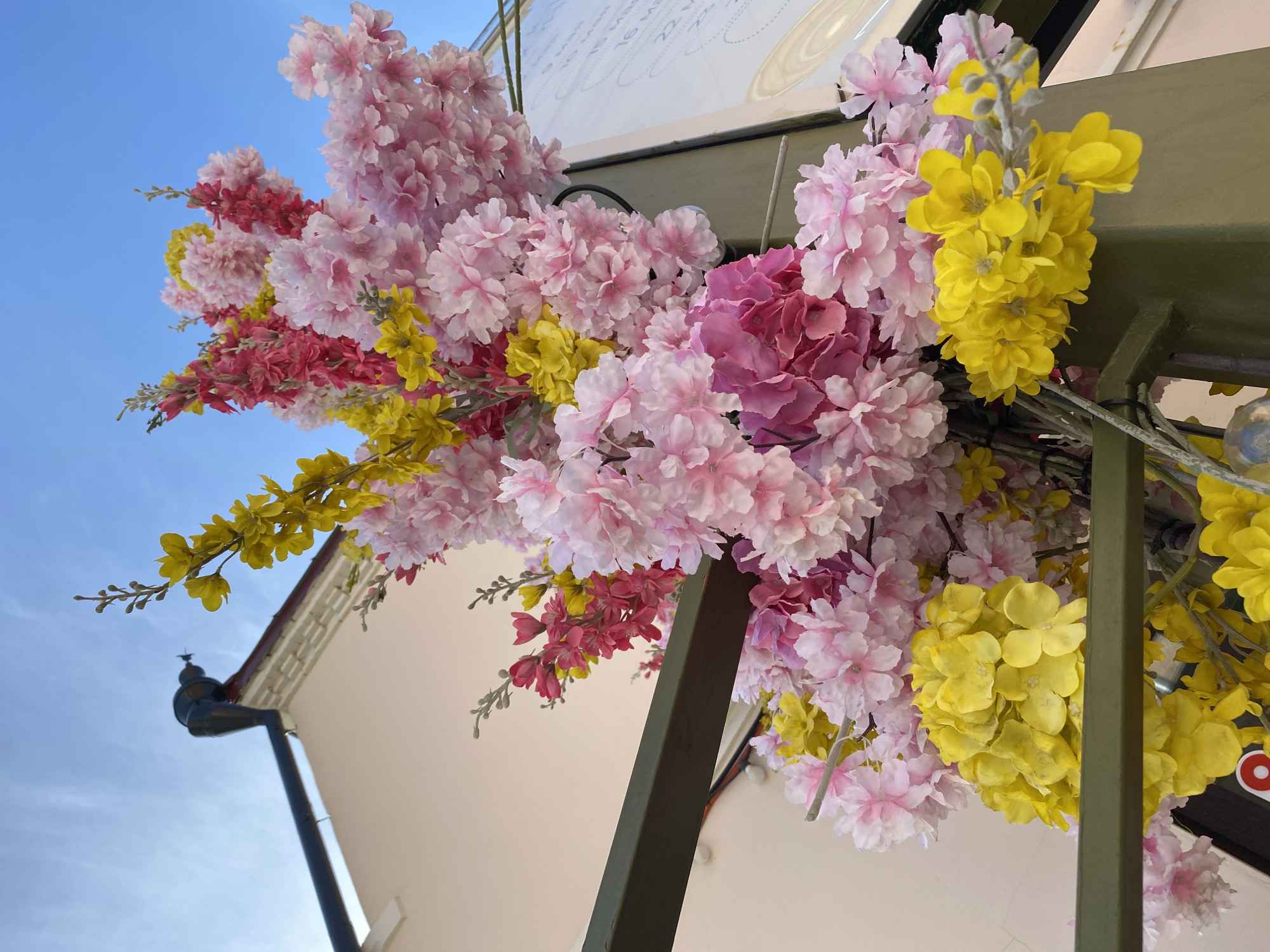 The image shows an outdoor display of pink, red, and yellow artificial flowers attached to a green metal frame. The frame is located next to a light-colored building with a dark-colored gutter system, with the blue sky visible in the background. The building is possibly in Izhevsk, Russia.