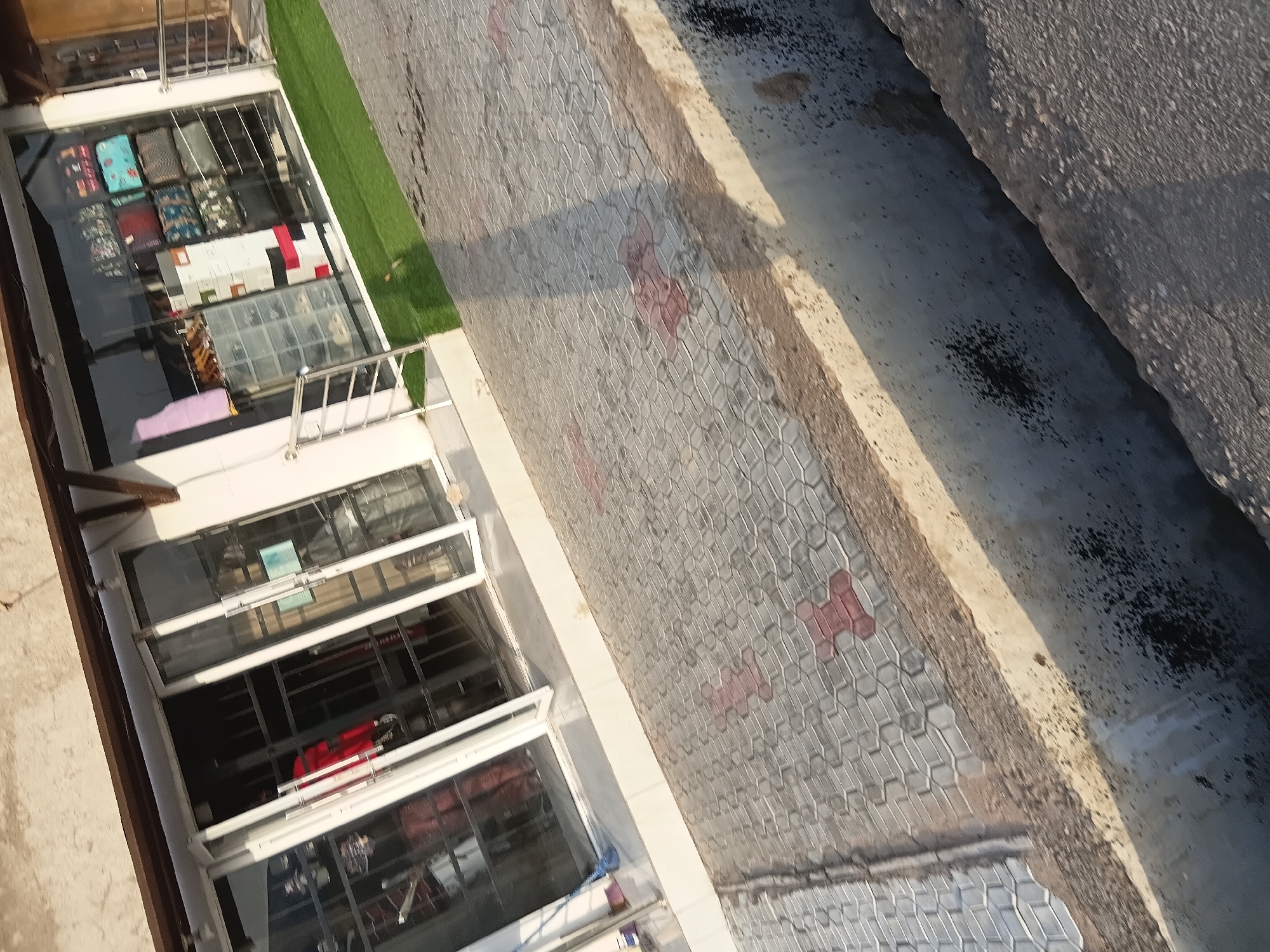 The image captures the exterior of a store located in Jalingo, Nigeria. The store features a large glass storefront with multiple panes, some of which are open, revealing an interior display of merchandise. Inside, racks and shelves are visible, holding various items, including clothing and accessories such as handbags. The floor outside the store is paved with interlocking gray tiles, interspersed with red hexagonal accent tiles. A strip of artificial turf runs along the edge of the storefront. In the foreground, a concrete structure with a rough texture and visible signs of wear and tear creates a raised border. The overall scene suggests a commercial area in a city.