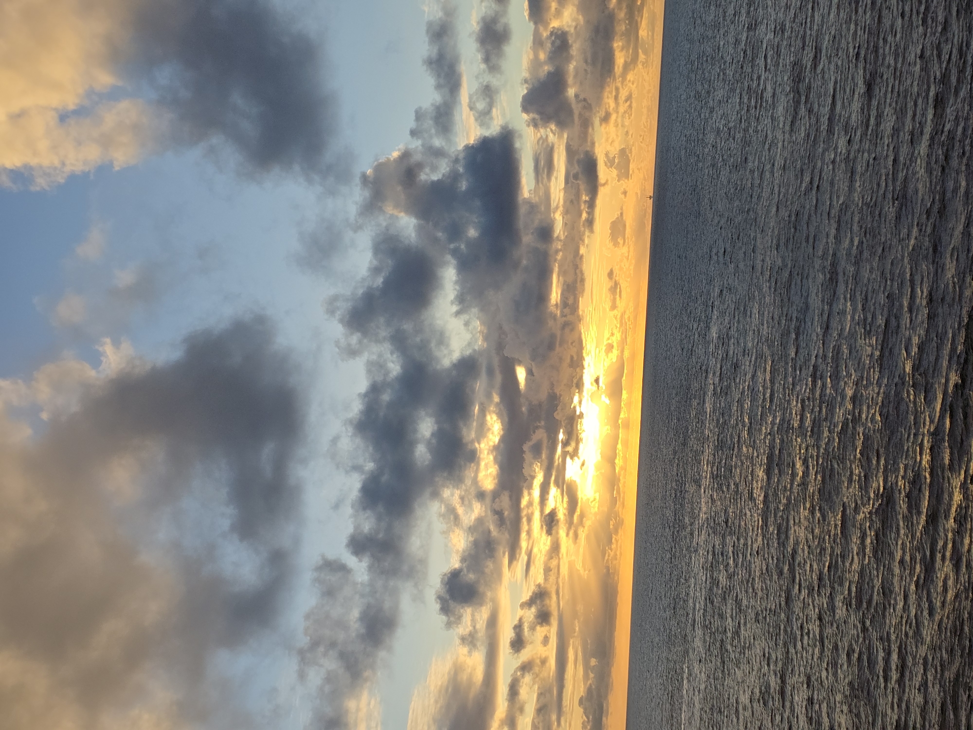 The image depicts a sunset over the ocean. The sky is filled with clouds, some dark and gray, others illuminated with golden hues from the setting sun. The ocean appears calm with subtle ripples reflecting the light. The location is unknown.