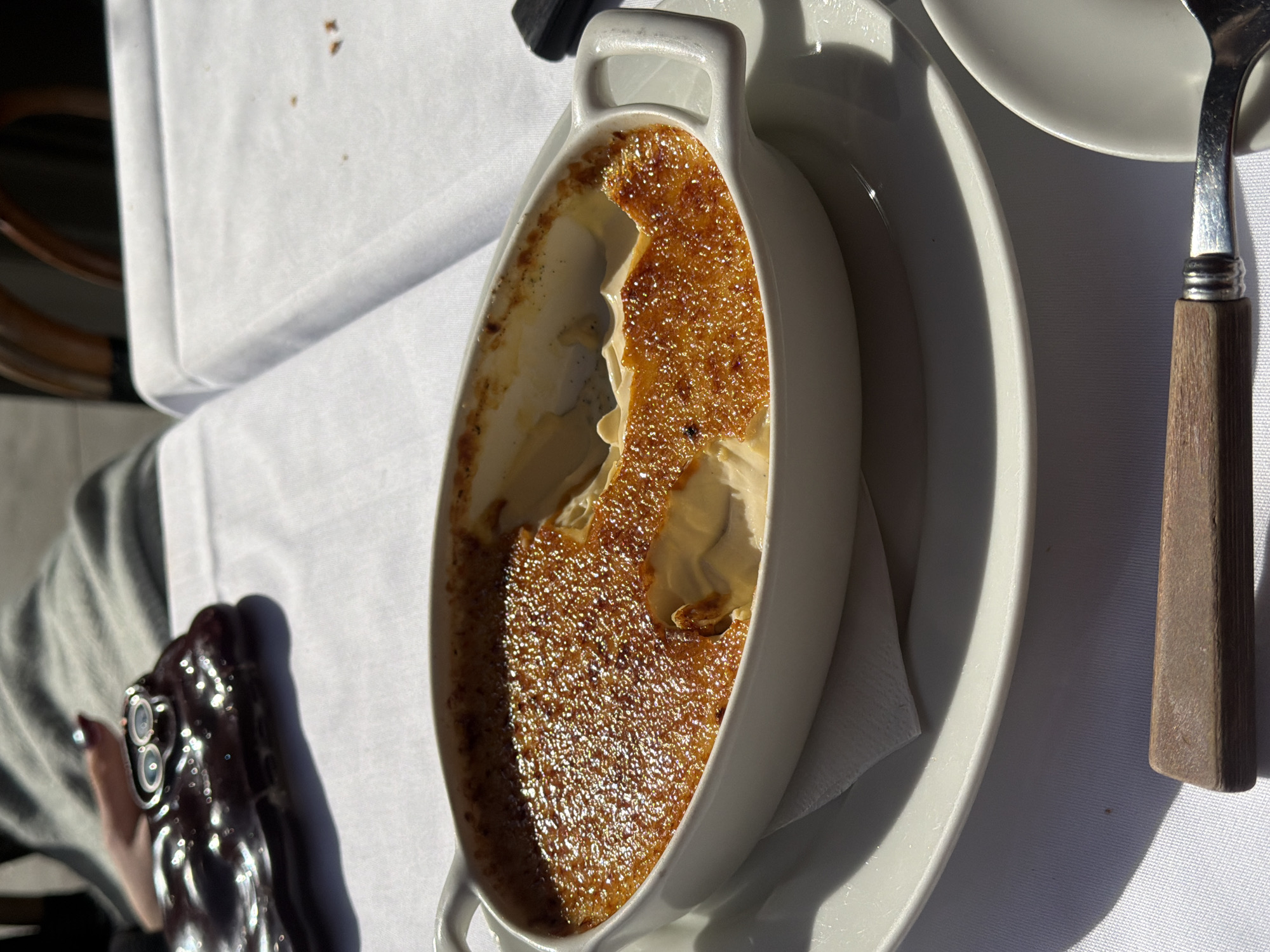 A partially eaten crème brûlée in a white ramekin is presented on a white plate with a folded white napkin. A spoon with a brown wooden handle rests to the right of the plate. A person wearing a gray shirt is seen in the lower left corner, holding a dark-colored smartphone with multiple camera lenses. The scene is set on a white tablecloth in a brightly lit environment. This is likely an indoor setting in London, United Kingdom.