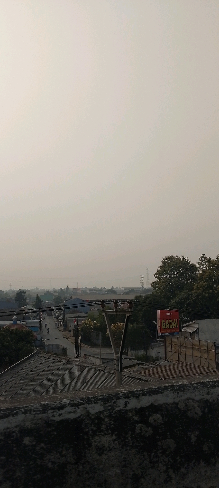 The image depicts a hazy, overcast day in an urban or semi-urban area of Bekasi, Indonesia. The foreground features a rough, textured concrete ledge, likely part of a rooftop or elevated structure. Behind this, corrugated metal roofs of several buildings are visible.
In the mid-ground, a road winds through the scene, with a few motorcycles and pedestrians on it. Buildings of various sizes and architectural styles line the road, some appearing residential and others more commercial or industrial. One building displays a red and white billboard with the text "KGI GADAI" and smaller text beneath it.
The background is dominated by a hazy sky, obscuring distant details. Several power line pylons are scattered across the landscape, indicating infrastructure in the area. Green trees are interspersed among the buildings, suggesting pockets of vegetation within the urban sprawl. The overall atmosphere is muted due to the haze, creating a somewhat subdued visual quality.