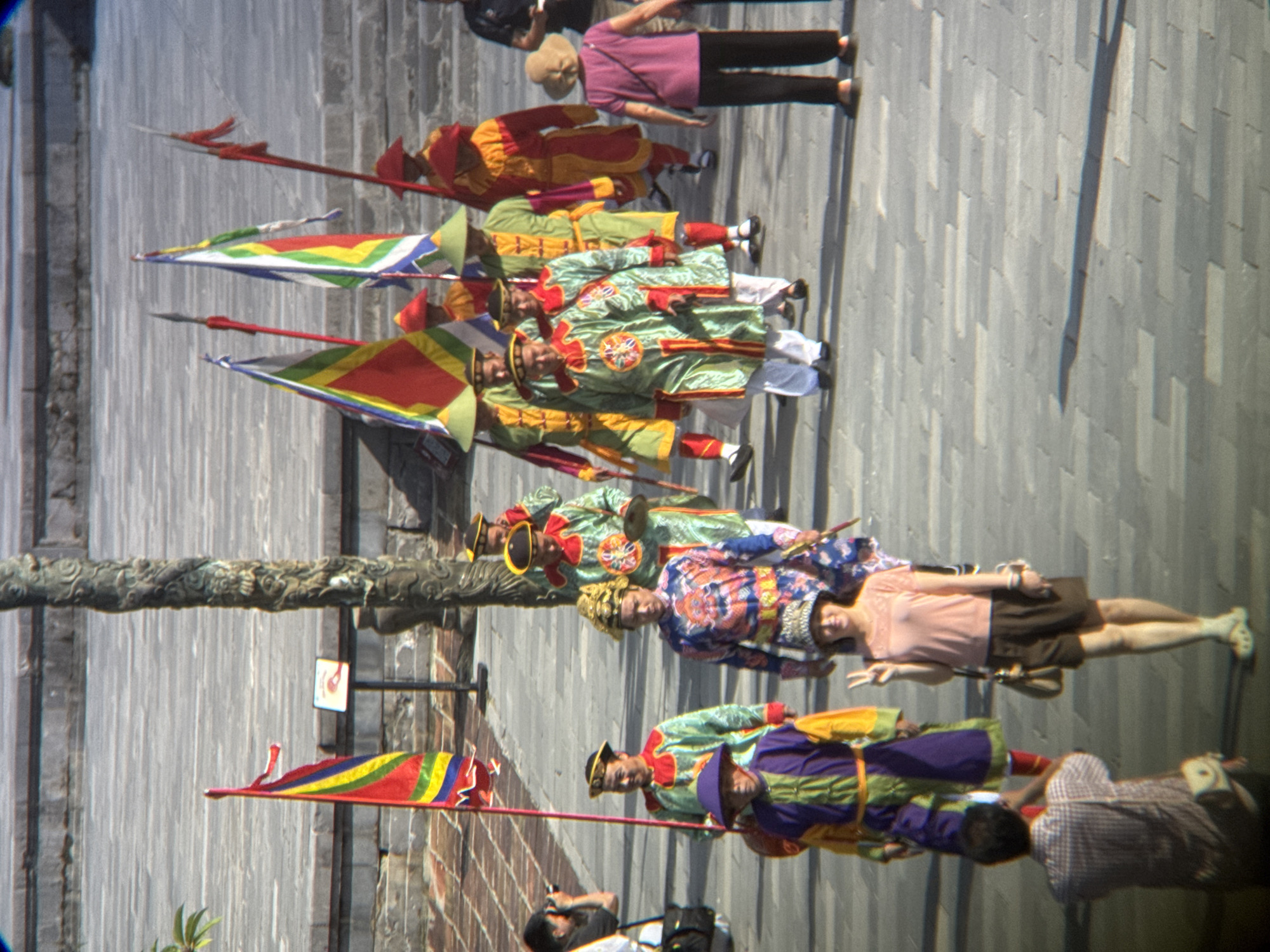 The image depicts a sunny outdoor scene on a paved plaza, likely in Phú Xuân Ward, Vietnam, based on the context provided. A group of individuals in traditional ceremonial attire, characterized by vibrant colors and ornate designs, are present. Several individuals are holding large, triangular flags on poles, with one flag prominently displaying red, yellow, and blue stripes. The people are adorned in long robes and conical hats, with some wearing elaborate headwear.
In the foreground, a woman with a light-colored hat and top is posing for a photograph, holding up two fingers in a peace sign. To her left, a man wearing a checkered shirt is taking a photograph of her with a camera. Further back, a line of men in ceremonial dress walks across the plaza. One man in the background is visible holding a camera, suggesting a public event or cultural demonstration. The environment features stone steps and a large stone pillar, indicating a historical or significant location. The ground is paved with rectangular stones.