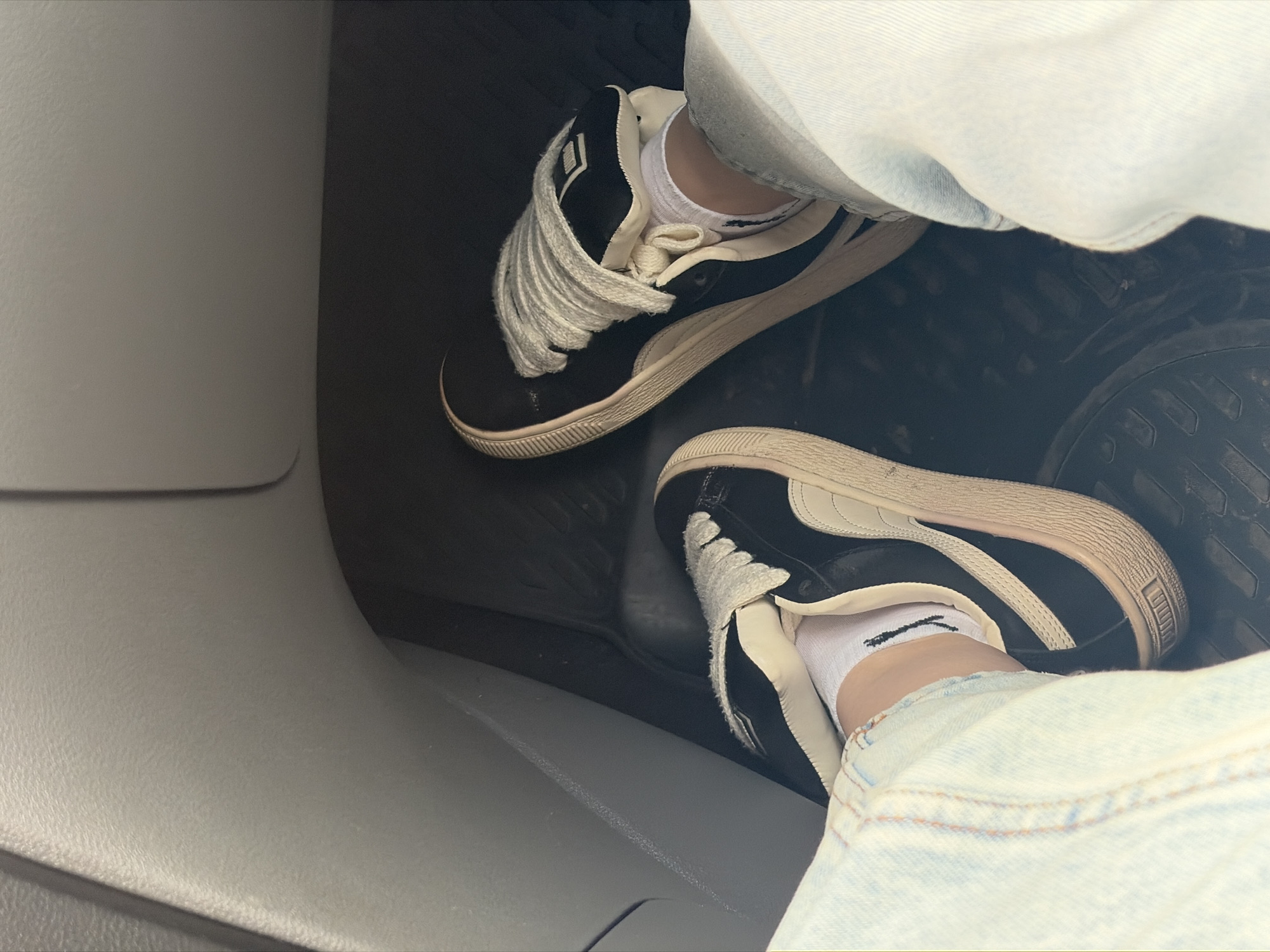 The image shows a pair of legs in light-wash denim jeans and white socks, wearing black and white Puma sneakers, sitting in the footwell of a vehicle. The interior of the vehicle is dark, and the trim is light grey. The location is assumed to be a car in the city of Chaikovsky, Russia, but that is not explicitly discernible from the image.