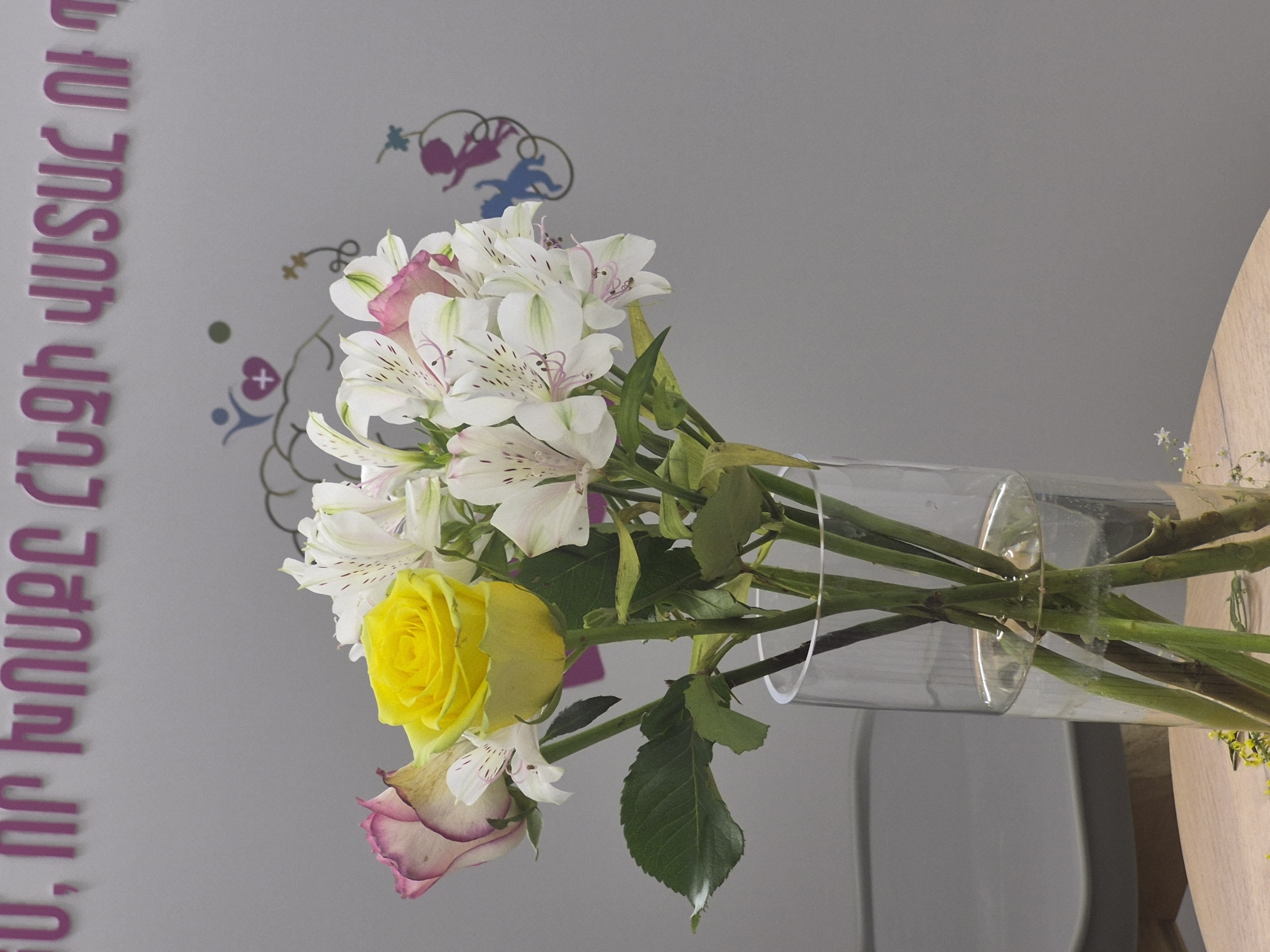 A vase of flowers sits on a table, with a blurred background featuring writing on a wall. The flowers consist of a bright yellow rose and several white alstroemerias with pink and green accents. The vase is clear glass, filled with water, and the stems of the flowers are visible within. In the background, large, capitalized letters in a pinkish-purple hue are displayed on a light gray wall, their text is illegible. To the right of the text, there are subtle, abstract graphic elements, including a silhouette of a person reaching towards a heart with a plus sign, and another stylized figure of a child. The overall setting appears to be indoors, possibly a public space or a venue in Yerevan, Armenia, given the presence of Armenian script on the wall. The lighting suggests an indoor environment, casting soft shadows.