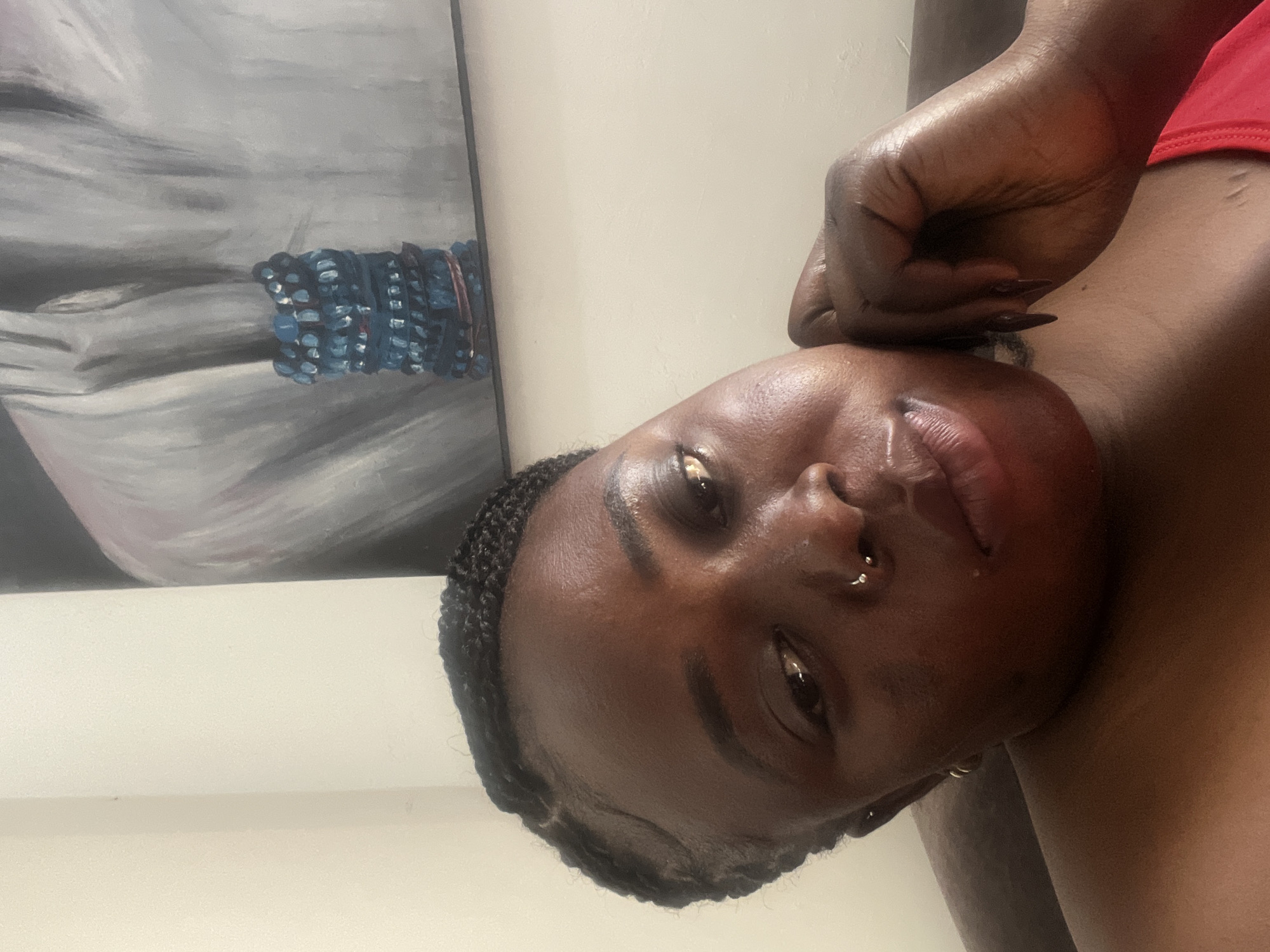 A selfie shows a Black woman with braided hair and a nose ring. She is leaning her head on her hand and looking at the camera. She is wearing a red top. Behind her is a painting of a person wearing a blue bracelet. The image appears to be taken indoors in a room with light-colored walls.