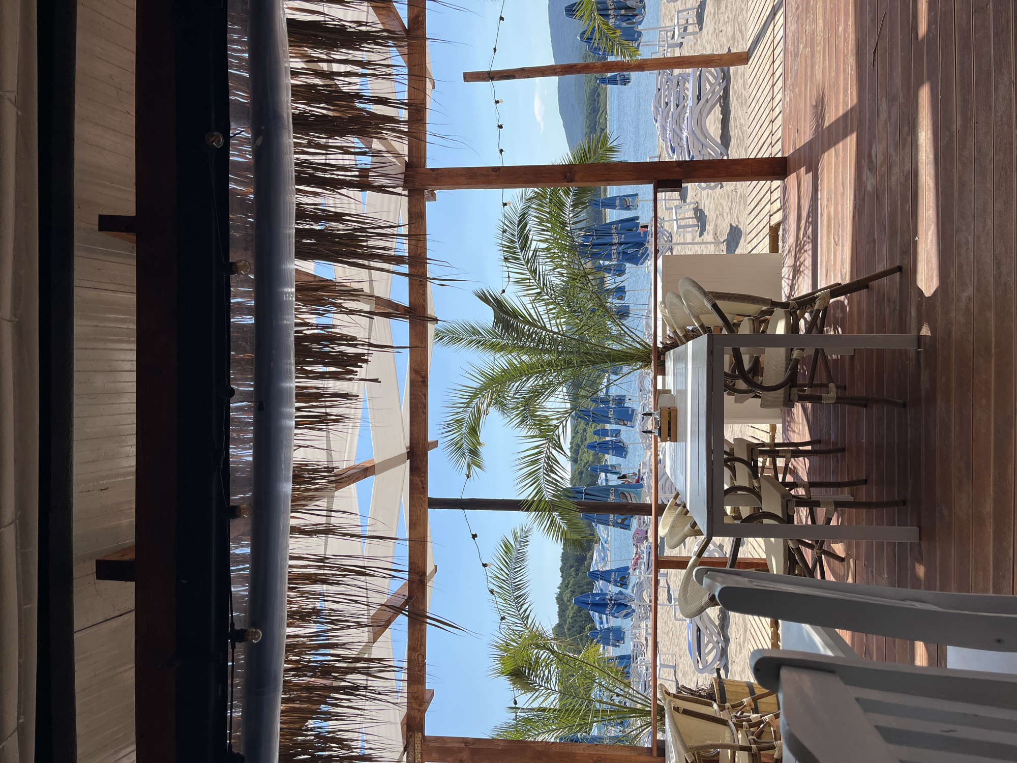 This media file depicts a serene beachside cafe scene in Ahtopol, Bulgaria. The foreground is dominated by a thatched roof structure supported by wooden beams, with white fabric awnings providing shade. Palm fronds are visible, adding to the tropical ambiance. Several tables and chairs, some made of wicker and wood, are arranged on a wooden deck.
Beyond the cafe, the scene opens up to a sandy beach dotted with numerous white lounge chairs and closed blue beach umbrellas. The calm, clear blue sea stretches towards a distant treed shoreline and hills under a bright, sunny sky, suggesting it is daytime, likely late morning or early afternoon, with clear weather. A string of small lights is strung across the wooden beams above. The overall impression is one of tranquility and relaxation, characteristic of a beach resort. No people are clearly visible or engaged in any specific activity, and no text is discernable in the image.
