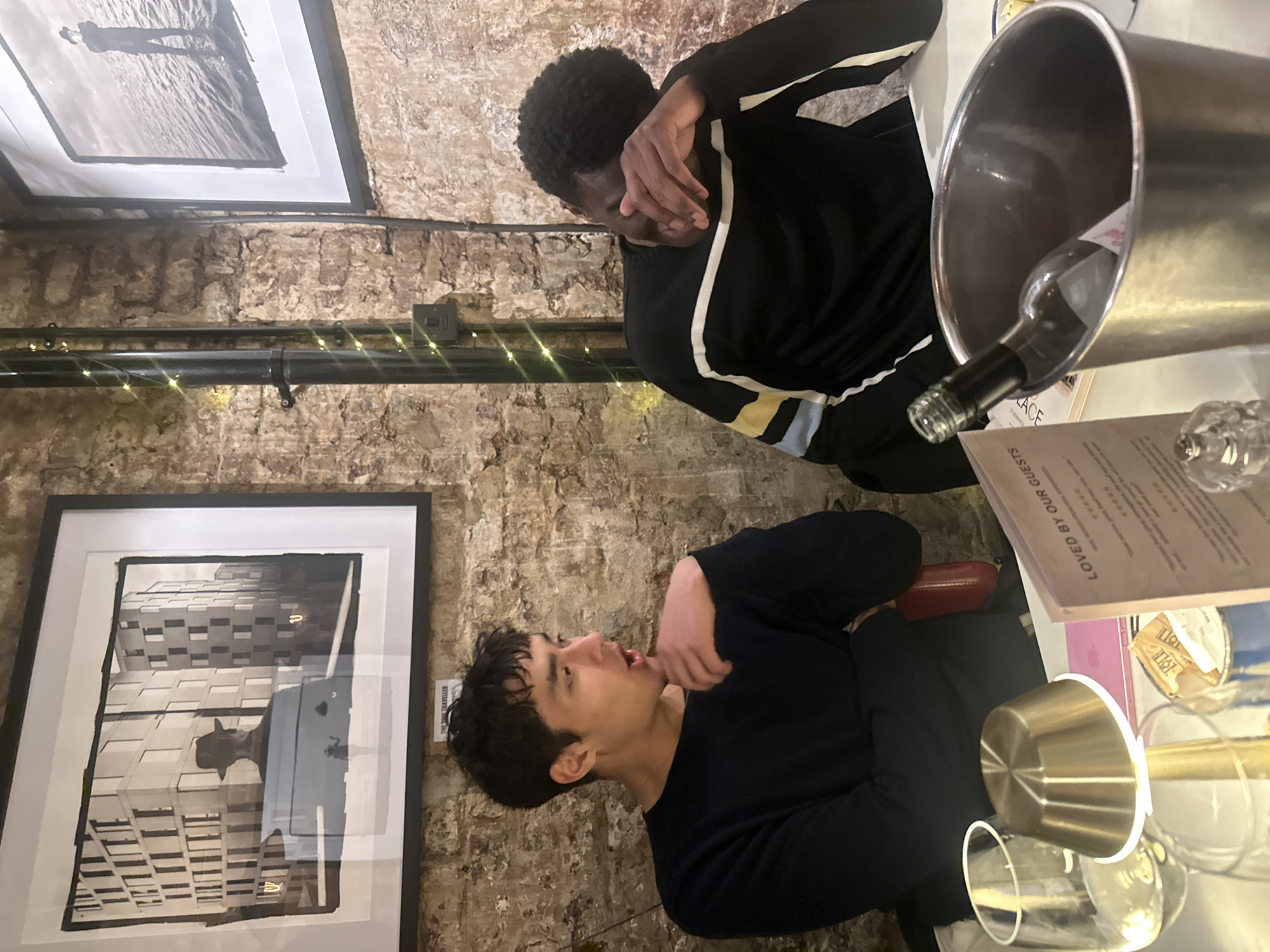 Two young men are seated at a table in what appears to be an indoor restaurant or bar setting in London. The background features exposed brick walls, contributing to a rustic ambiance, illuminated by warm string lights wrapped around a black pipe running across the wall.
The man in the foreground, with short dark hair and wearing a dark long-sleeved top, is looking upwards with an open-mouthed, expressive gaze, his right hand near his chin. The man in the background, also with short dark hair and wearing a black striped long-sleeved top, is leaning forward with his face partially covered by his hand, possibly in a gesture of shyness, amusement, or reflection.
On the table, a silver ice bucket holds a wine bottle, alongside an empty wine glass and a small metallic cup. A menu card on the table clearly reads "LOVED BY OUR GUESTS." Two framed black and white artworks hang on the brick wall: the top one shows a person standing in shallow water, and the bottom one depicts a building facade with a silhouette in the foreground. A small white tag next to the bottom artwork bears the text "THE TIME TRAVELLER." The scene suggests an evening gathering, possibly over drinks and conversation, in a cozy and artfully decorated space.