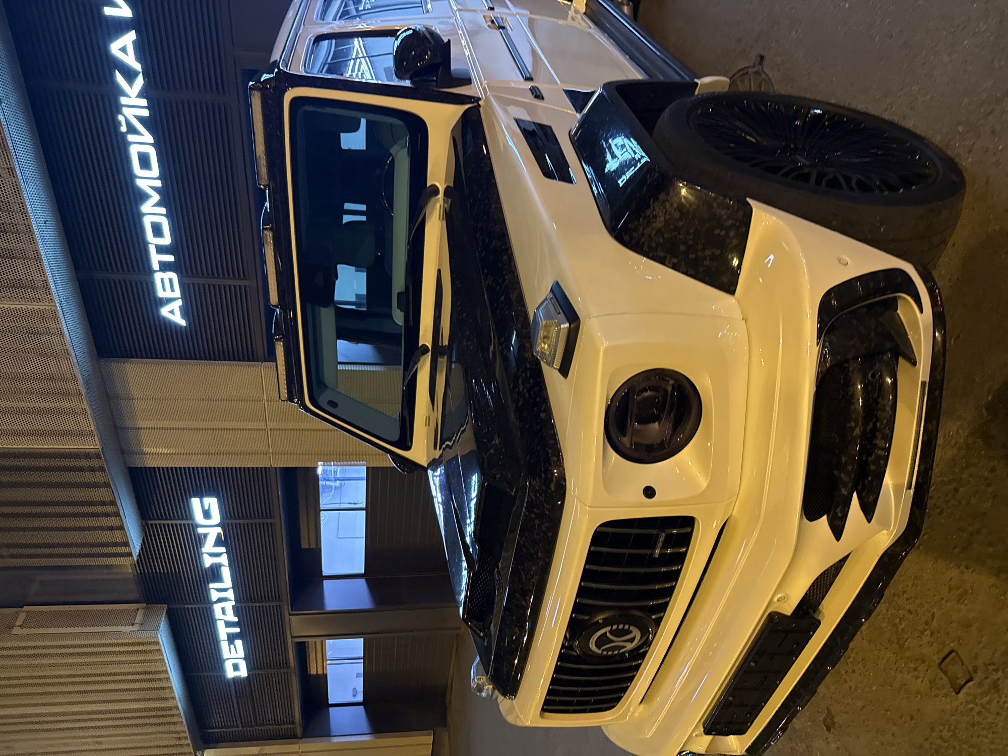 The image depicts a white Mercedes-Benz G-Class SUV with aftermarket carbon fiber body panels, parked outside a building displaying illuminated signs in Russian Cyrillic reading "АВТОМОЙКА" (car wash) and "DETAILING." The vehicle is situated on a paved surface, likely in an urban setting. The photograph was taken at night, and the perspective is from a low angle, highlighting the vehicle's front and side profile. The location appears to be in Moscow, Russia, based on the Cyrillic signage.