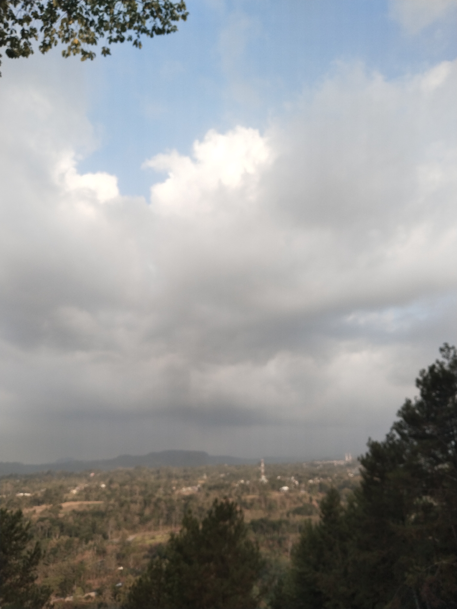 The image depicts an overcast sky with patches of blue visible, hinting at the remnants of sunlight. In the foreground, the tops of trees with sparse foliage are visible, with one branch extending from the upper left corner. The mid-ground presents a vast, blurred landscape that appears to be a city or town, identified as Sokatengah, Indonesia. Buildings, including what might be a water tower and other structures, are scattered across the terrain, surrounded by a dense arrangement of trees. The background is dominated by rolling hills or mountains, their forms softened by the atmospheric conditions. The overall impression is one of a panoramic view of a semi-urbanized natural environment under a cloudy sky.