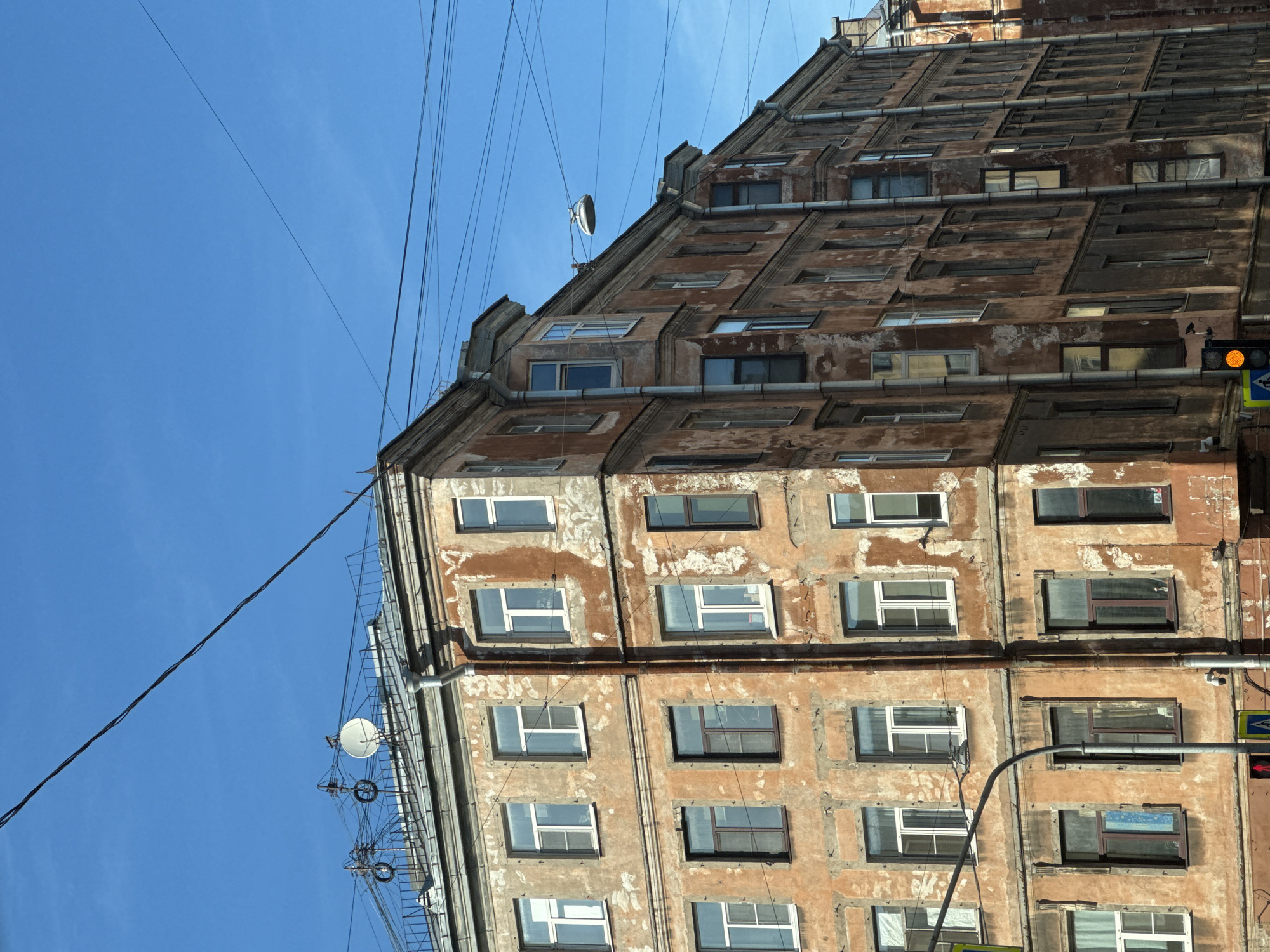 This is a medium shot of an old, weathered, five-story apartment building with a reddish-orange facade in Saint Petersburg, Russia. The building has a corner section that juts out, and multiple windows are visible on each floor, many with white frames and some with darker frames. The paint on the building is peeling and cracked, revealing a lighter colored plaster underneath. Several vertical drainpipes run down the side of the building.
The building is situated on a clear day with a bright blue sky. Overhead, multiple power lines and communication cables crisscross the sky. On the roof of the building, there are several antennas and a satellite dish. A street lamp is mounted on a pole extending from the building's upper facade.
At the bottom of the image, the edge of a sidewalk is visible, along with traffic signals and pedestrian crossing signs. A yellow traffic light is illuminated. The overall impression is of an aged urban environment in Saint Petersburg.