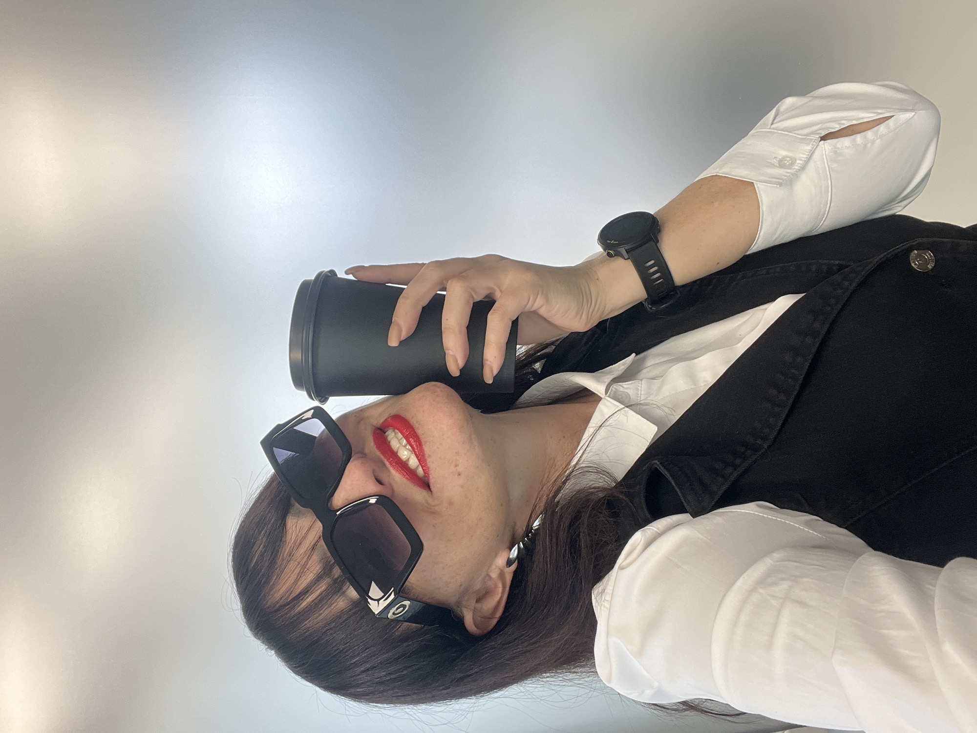 The image shows a woman wearing black sunglasses, red lipstick, a white shirt, and a black vest. She has a black watch on her left wrist and is holding a black cylindrical object, likely a coffee cup or tumbler. The background is a neutral gradient of white to light gray. The location in the city of Perm, Russia, is not discernible from the image.