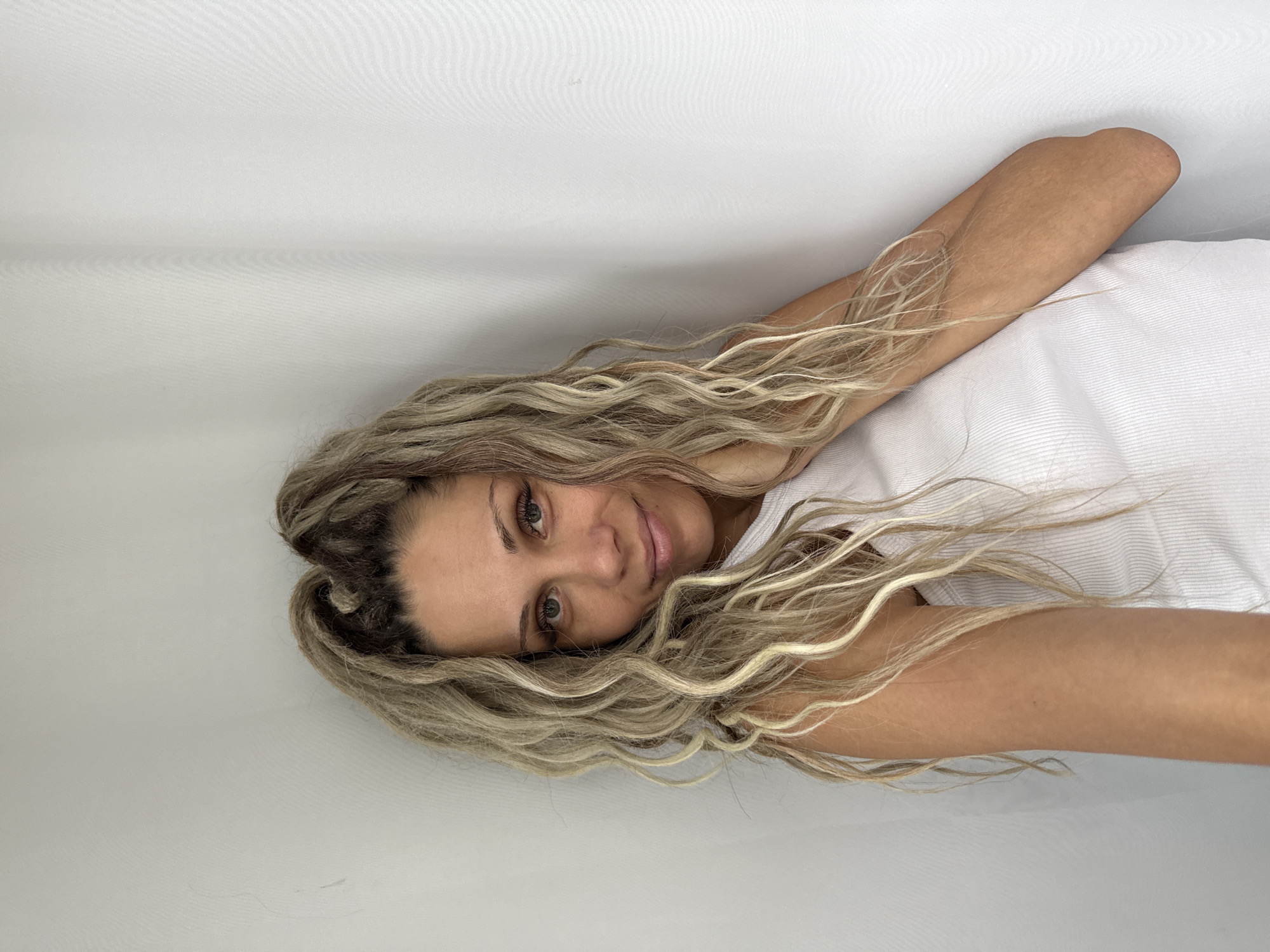 A woman with blonde, curly dreadlocks is positioned in the corner of a room, leaning her head against the wall. She is wearing a white tank top. Her right arm is bent, with her hand resting on her neck and shoulder. Her left arm is not fully visible. The background consists of two plain white walls, with a textured, slightly wrinkled white curtain to the right. The lighting is bright and diffused. There are no discernable elements to indicate a specific location within the city of Rusanovo, Russia.