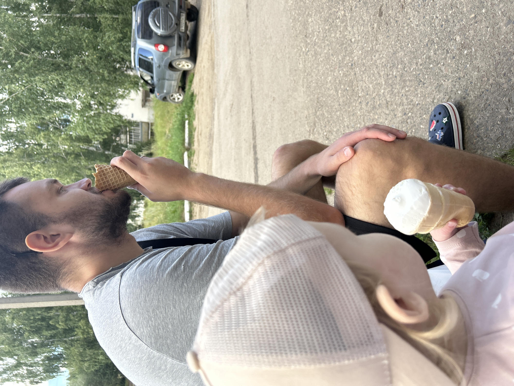 The image depicts two people, a man and a young child, sitting outdoors and eating ice cream. The man, on the right, is wearing a grey t-shirt and has a beard, and is holding an ice cream cone to his mouth. The child, on the left, is wearing a pink long-sleeved shirt and a light-colored cap, and is holding an ice cream cone. They are sitting on a paved surface, possibly a sidewalk or a pathway. In the background, there are trees, a building, and a dark-colored SUV. The overall setting appears to be a residential area on a sunny day. The specific location is stated to be Noviy, Russia, though no explicit markers within the image confirm this.