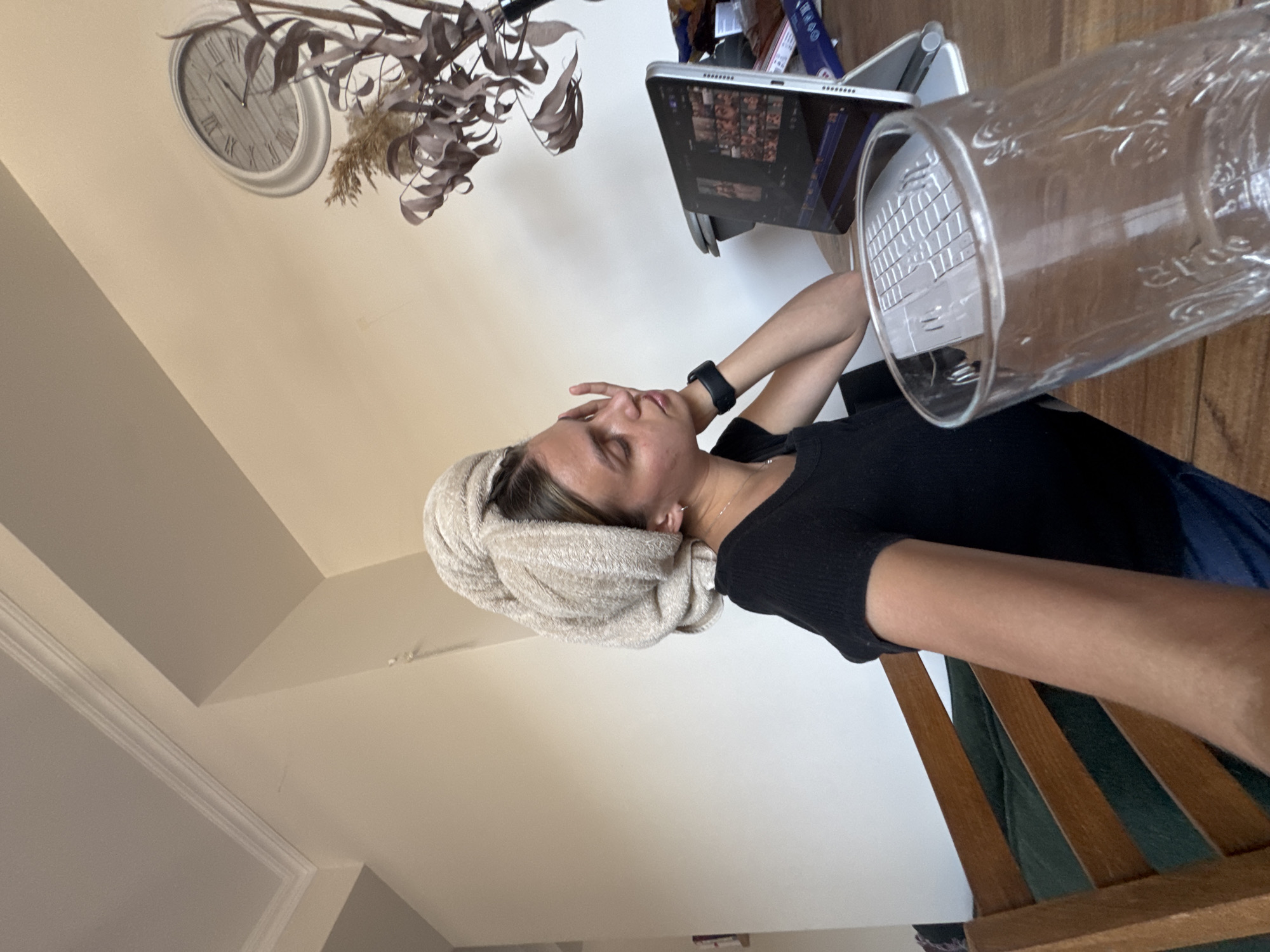 The image captures an indoor scene featuring a young woman who appears to be resting or relaxing. Her hair is wrapped in a light-colored towel, suggesting she may have recently showered. She is wearing a dark, possibly black, short-sleeved top and her left arm, adorned with a smart watch, is raised with her hand touching her forehead or face. Her eyes are closed or partially closed, conveying a sense of weariness or peaceful contemplation.
To her right, a laptop or tablet is open on a wooden surface, its screen displaying a grid of small images or thumbnails. A transparent glass or cup, possibly with decorative embossing or text that is not clearly legible, is prominently positioned in the foreground, close to the camera, with the reflection or view of the laptop's keyboard visible through it.
In the background, to the left, a wall clock with Roman numerals (I through XII) is visible. The clock's hands are positioned around the XI and II marks, making the exact time ambiguous. Dried plant decorations hang near the clock. The walls are a light, neutral color, and crown molding is visible in some areas, indicating a structured interior space, likely a home living room or bedroom. The woman is seated on a chair with visible wooden slats. The overall atmosphere suggests a quiet, personal moment of rest.