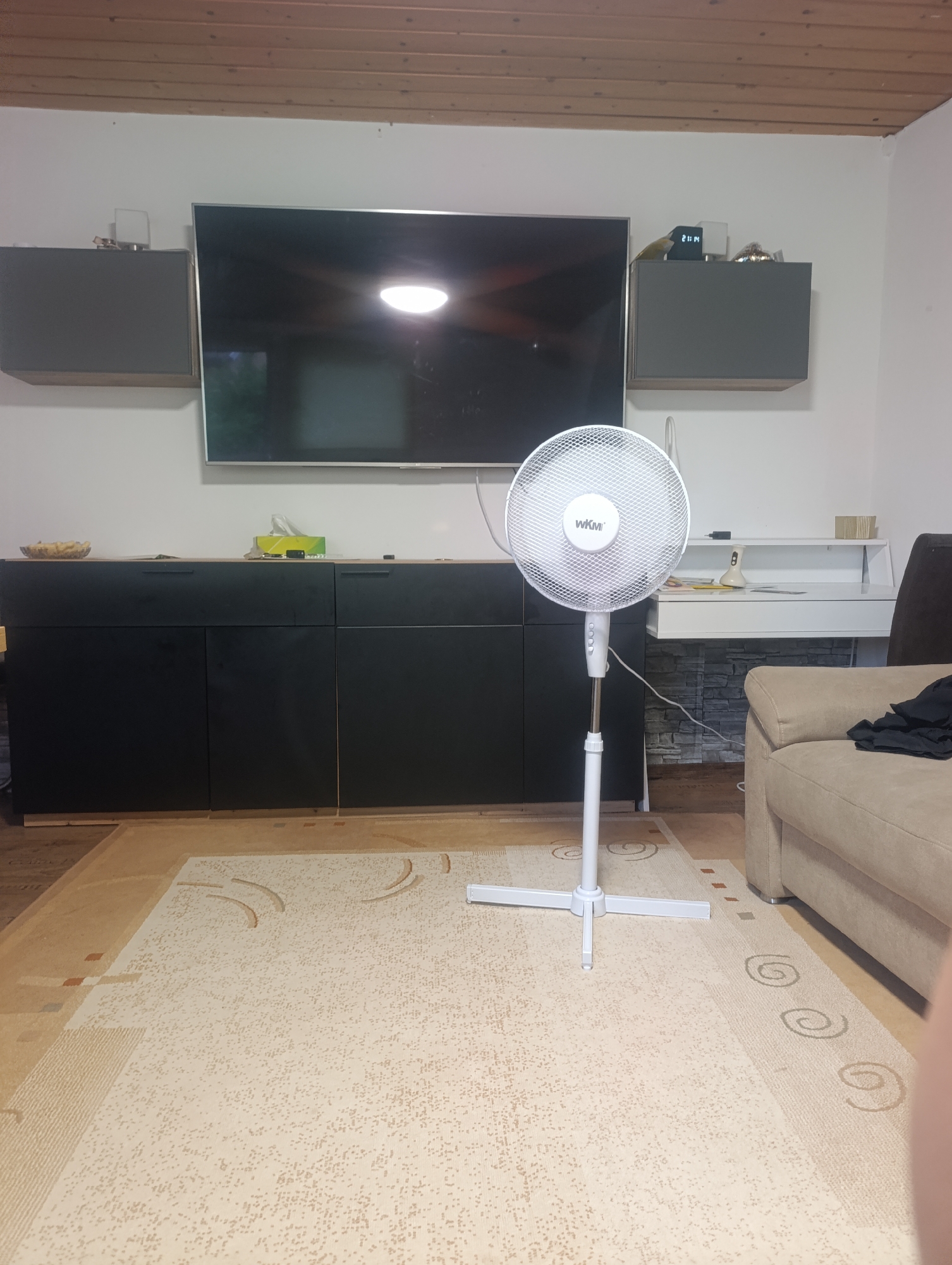 The image depicts an interior scene of a room in Pfullingen, Germany. The room is furnished with a large flat-screen television mounted on a white wall, flanked by two gray wall-mounted cabinets. Below the television is a black console cabinet with a light-colored countertop, upon which are various items including a bowl, tissues, and other indiscernible objects.
To the right of the console is a white desk with a lamp and other items, set against a background of what appears to be faux stone or textured wall covering. Standing in the foreground is a white electric fan. The floor is covered with a light-colored patterned rug. A section of a beige sofa is visible on the right side of the image. The ceiling is wood paneled.