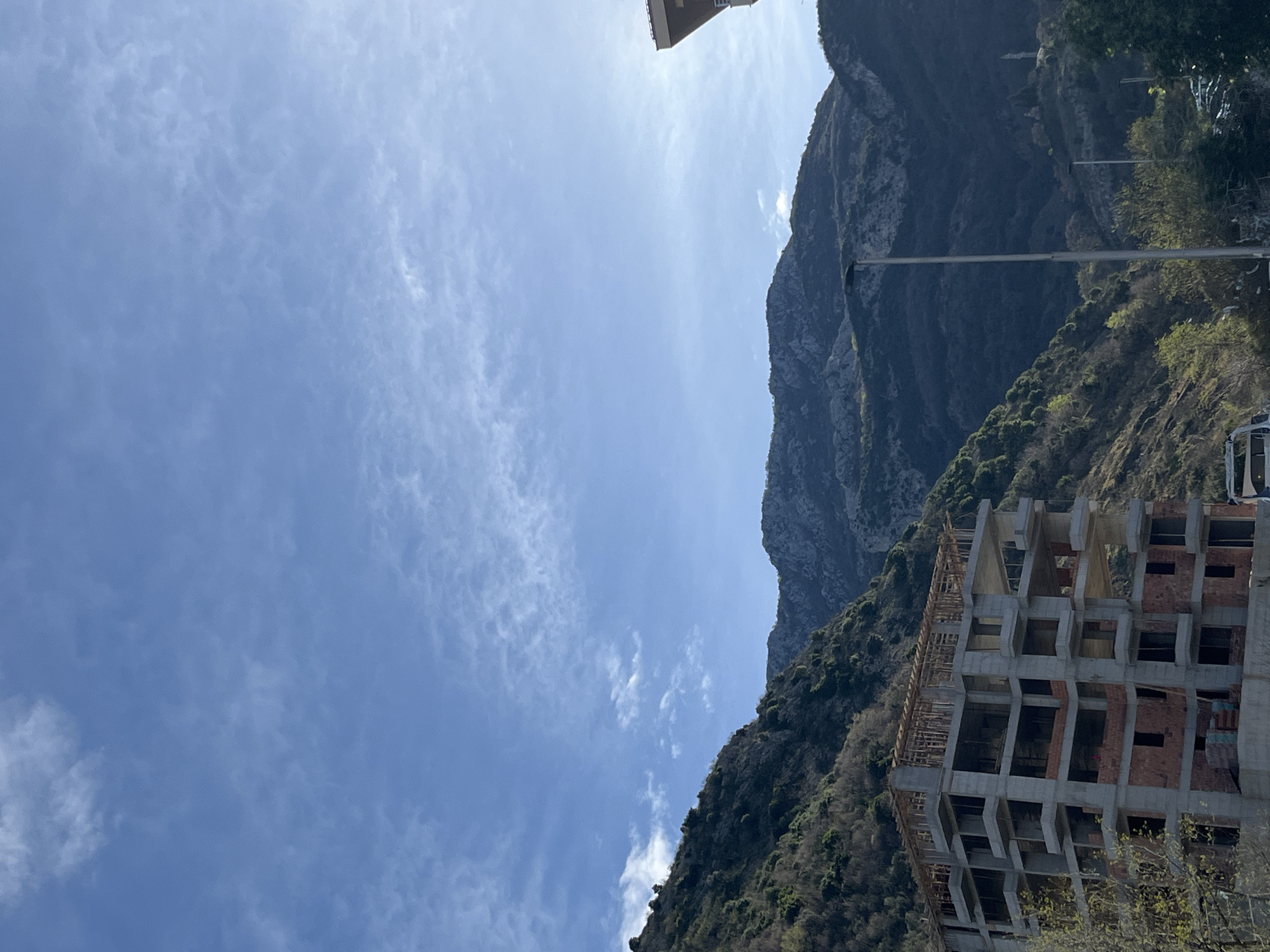 The image shows a partially constructed building located in Rafailovići, Montenegro. The building is a multi-story structure made of concrete and brick. It is situated on a hillside with trees and mountains in the background. The sky is blue with some clouds.