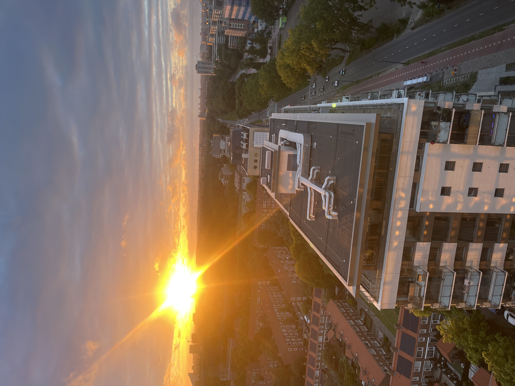 This image captures a vibrant sunset over a cityscape, likely taken from an elevated perspective, possibly a building. The sun, a brilliant burst of golden light, is positioned in the upper left quadrant of the sky, casting long rays across the horizon. The sky is a gradient of warm oranges, pinks, and purples, transitioning to a cooler blue-grey at the top, with scattered clouds tinged by the sunset's glow.
Below the sky, the cityscape unfolds. In the foreground, a modern apartment building with balconies and large windows dominates the right side of the frame. Its white and dark facade is illuminated by the sun's warm light, creating reflections. Adjacent to this building, a street runs parallel, lined with trees and a red-colored bike lane. Several cars are visible on the road.
Further into the distance, rows of residential buildings with red-tiled roofs are visible. These buildings appear to be of a lower density than the apartment building in the foreground. Beyond them, more buildings and trees extend towards the horizon, where a hazy silhouette of the city skyline can be seen against the bright sky. The overall scene conveys a sense of urban life at dusk, bathed in the dramatic light of a setting sun.
