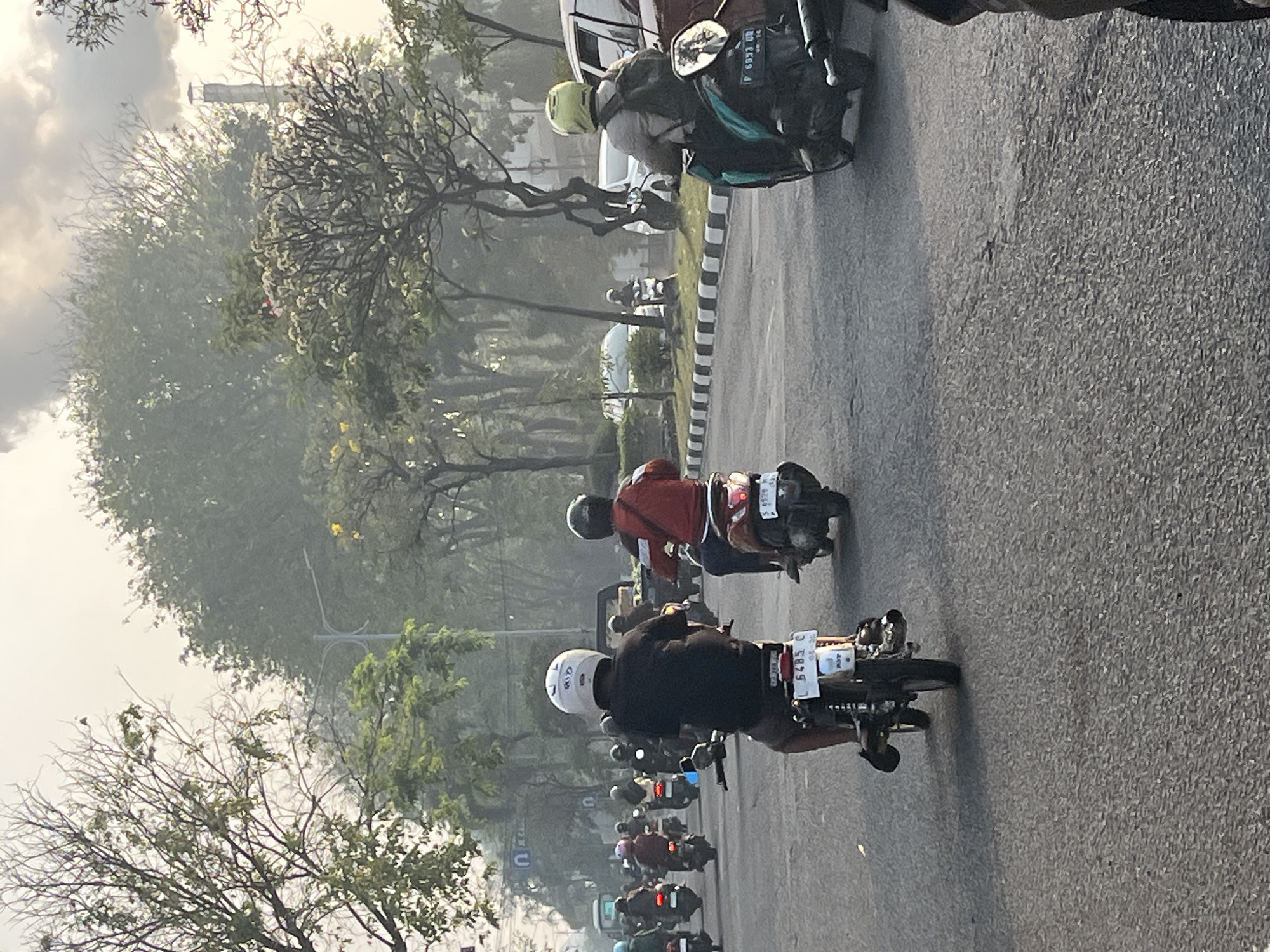 The image depicts a street scene in Candi, Indonesia, characterized by a hazy, overcast sky and lush greenery lining the road. A group of people on motorcycles are visible, navigating the asphalt street. In the foreground, a person wearing a black shirt and a white helmet rides a motorcycle with a white license plate. To the right, another person in an orange jacket and a black helmet is on a scooter with a visible license plate. Further down the street, several other motorcycles and vehicles are present, some with riders and some without. The environment appears to be a typical urban or suburban street with trees and possibly buildings in the background, partially obscured by the atmospheric conditions. The overall impression is one of daily commute or travel in a tropical environment.