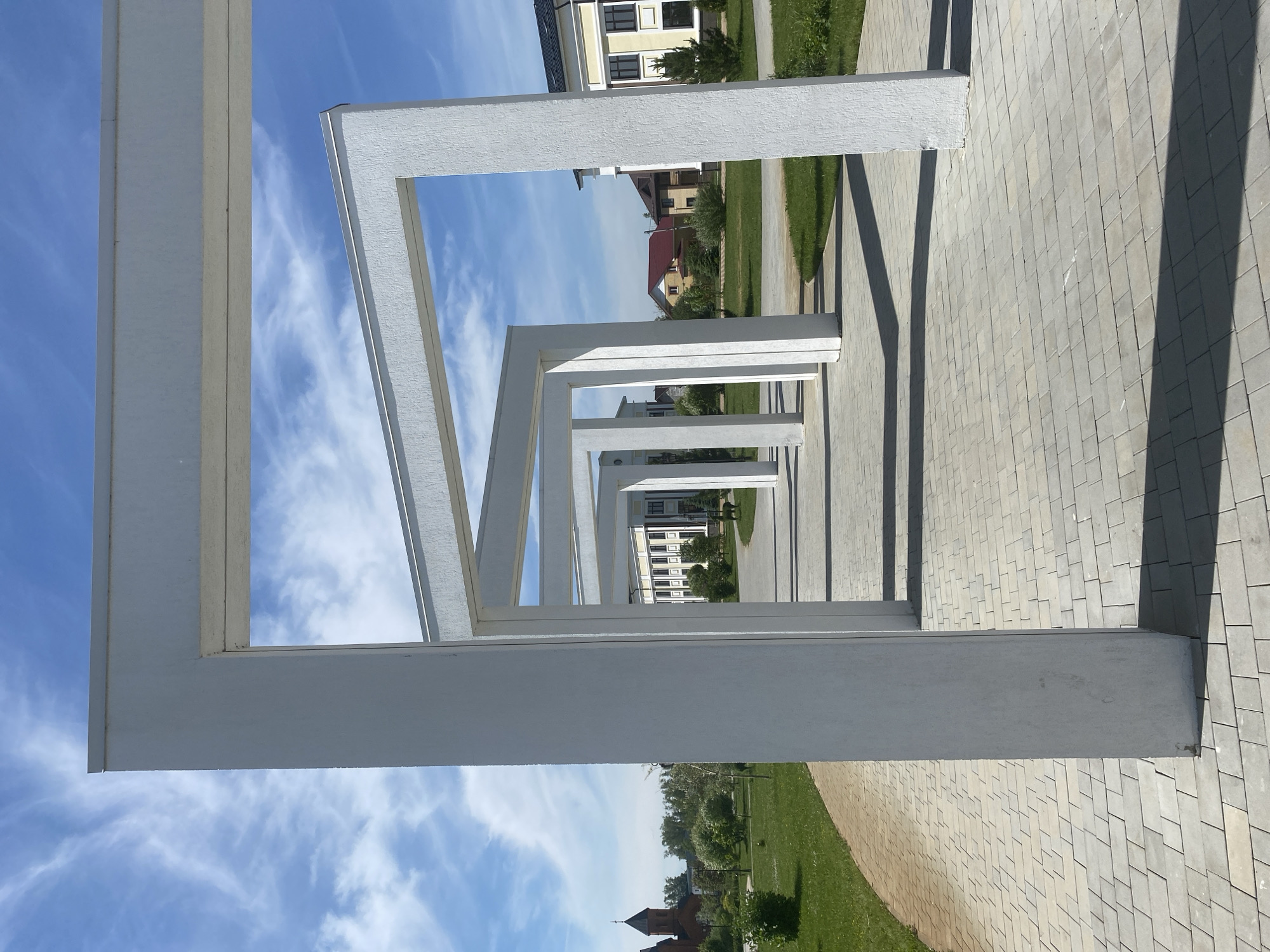 A vertically oriented photograph, taken in Tyumen, Russia, captures a modern outdoor walkway featuring a series of receding white architectural structures under a bright blue sky. The main subjects are multiple rectangular, arch-like frames, likely forming a pergola or a sculptural pathway, constructed from a white-painted material with a subtly textured finish. These structures are arranged linearly, creating strong leading lines and a sense of depth as they recede into the distance.
The ground beneath the structures is paved with light-grey rectangular interlocking pavers. To the left of the paved area, a strip of vibrant green grass is visible. Sunlight casts long, distinct shadows of the white structures onto the paved surface, indicating a clear, sunny day, likely in the morning or late afternoon. The sky above is a bright blue with scattered, wispy white clouds, suggesting fair weather conditions.
In the background, beyond the white framework, several buildings are visible. Some appear to be modern residential or commercial structures with light-colored facades and dark windows. Further in the distance, a building with a reddish-brown roof and spire-like elements can be seen, possibly a church or a traditional building, offering a contrast to the contemporary architecture. There are no people visible in the scene, and no discernible text. The overall impression is one of a clean, well-designed, and serene urban or park-like outdoor space.