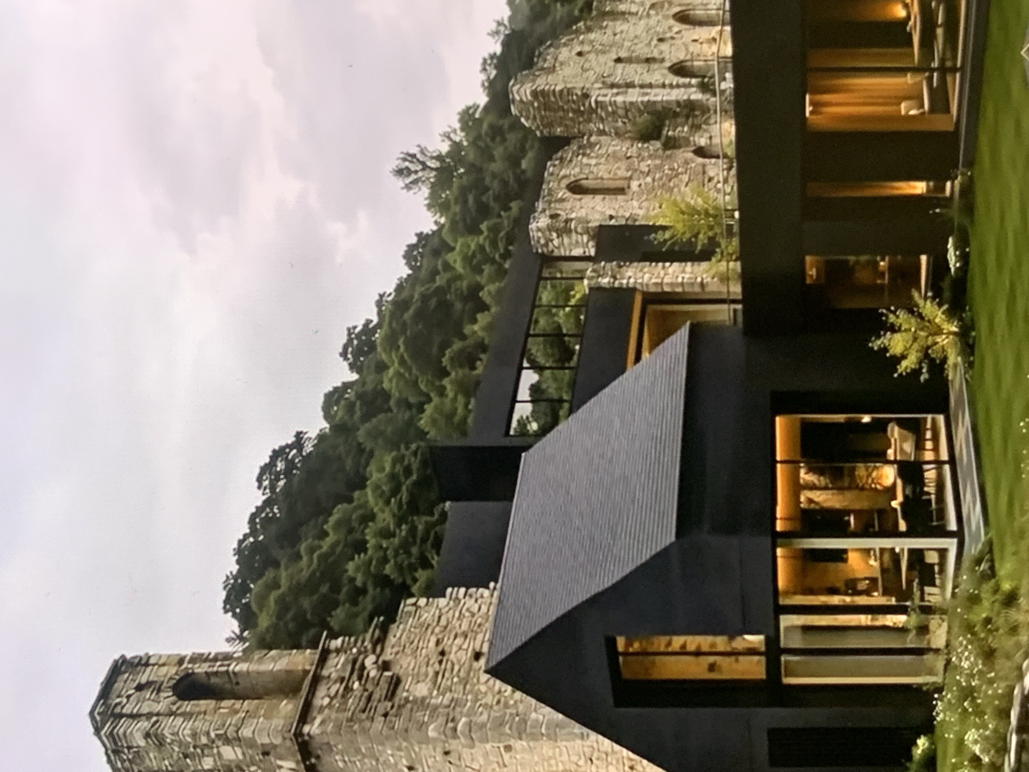 The image displays a modern black house nestled beside ancient stone ruins. The house features large glass windows that illuminate the interior with a warm, golden light, revealing glimpses of furniture and dining areas. The ruins, partially overgrown with greenery, stand on a hillside covered with dense trees. A stone tower, also part of the ruins, is visible to the left of the house. The sky is overcast with soft, diffused light. The scene is set outdoors, with a well-maintained lawn in the foreground. The image does not show any people. There is no indication that this location is in Stavropol, Russia.
