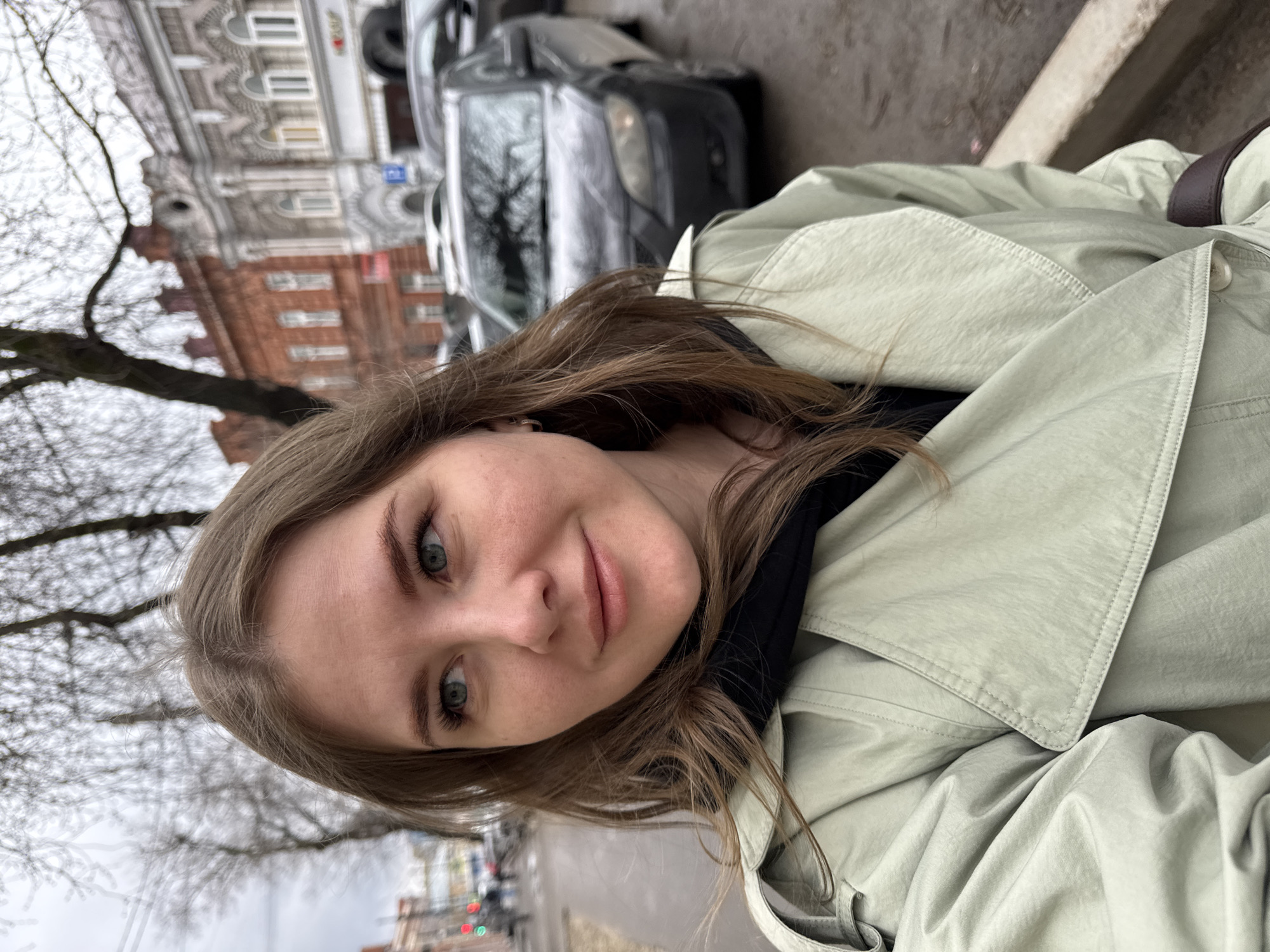A woman is taking a selfie in Perm, Russia. She is wearing a light green coat. Behind her, there is a dark-colored car, a building with intricate architecture, and bare trees. In the background, there is a traffic light and other vehicles. The sky appears overcast.