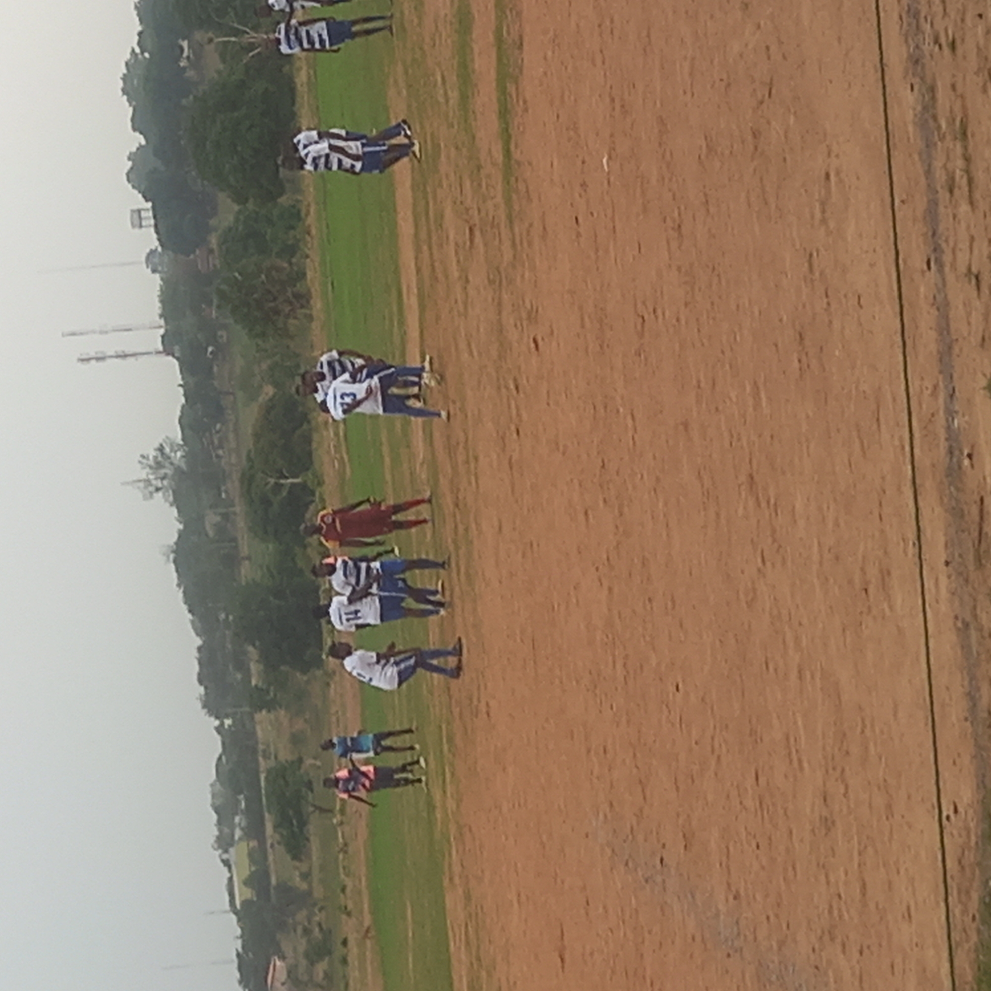 A football game is in progress on a grassy pitch adjacent to a dirt field. The location is Narayi, Nigeria. Several players are dressed in white and blue striped uniforms, while one is wearing red. The players are dispersed across the field. Beyond the pitch are trees and several communications towers.