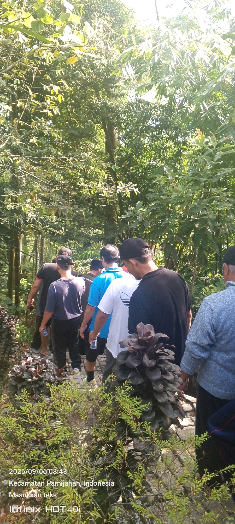 A group of men are walking on a paved path through a dense, sun-dappled forest. The path is lined with ornamental plants, some with dark purple foliage. The men are walking away from the camera in single file. They are dressed in casual clothing, including t-shirts, shorts, and pants. One man in the foreground wears a black t-shirt and a black baseball cap. To his left, another man wears a blue t-shirt and black shorts. Further down the path, a man in a dark t-shirt and black pants is visible. The forest canopy is thick with green leaves, and sunlight filters through, creating bright patches and shadows on the path. The date stamp in the lower left corner reads "2026/09/06 08:43" and the location is identified as "Kecamatan Pamijahan, Indonesia". A message "Masukkan teks" and "Infinix HOT 40i" are also visible.