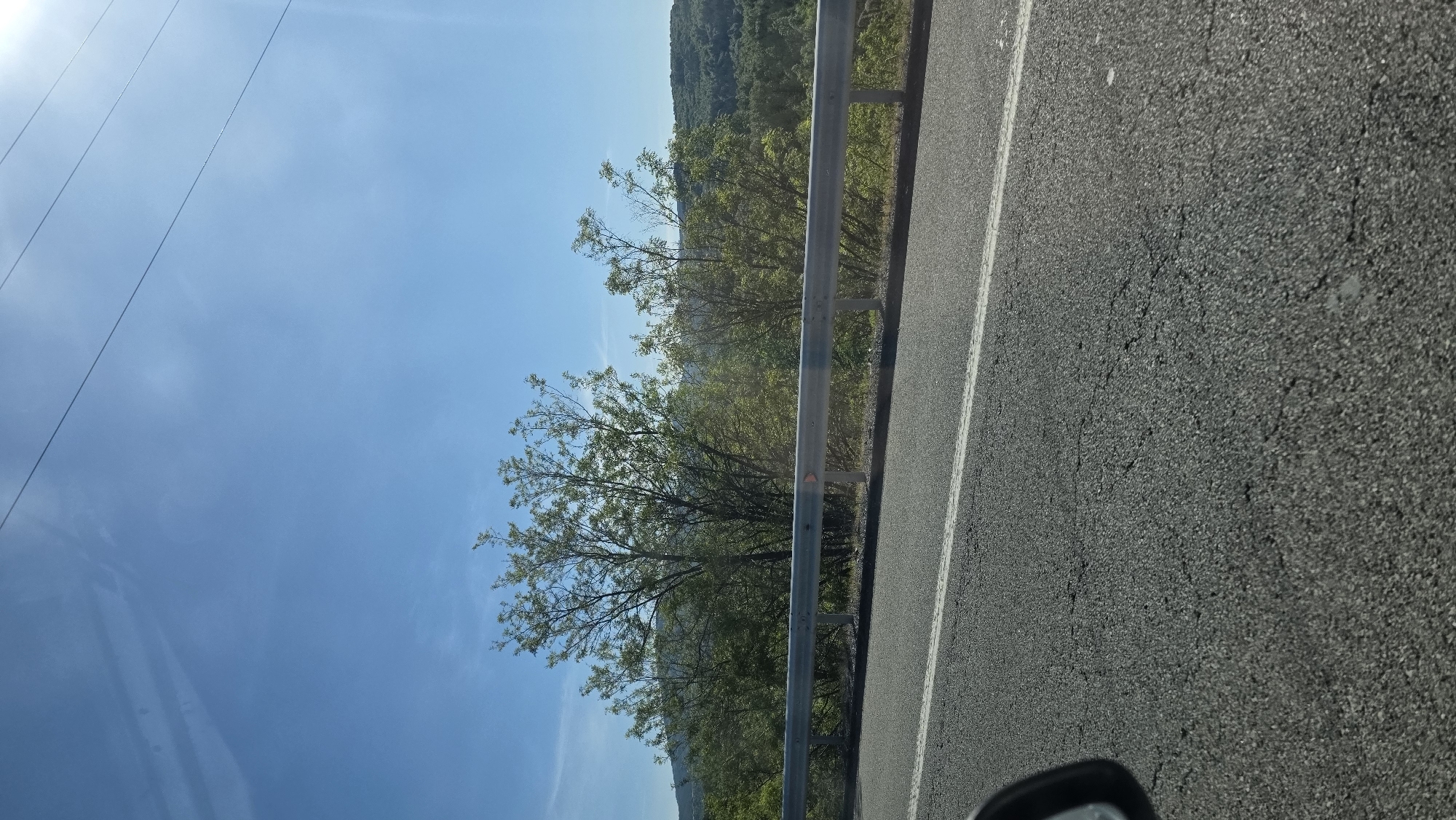 The image shows a view from a vehicle traveling along a road in Dyakovo, Bulgaria. To the right, the asphalt road is marked with a white line. A silver guardrail separates the road from a hillside with trees and vegetation. Above and to the left, the sky is visible. Power lines are also present. The overall scene appears to be captured during daylight hours.