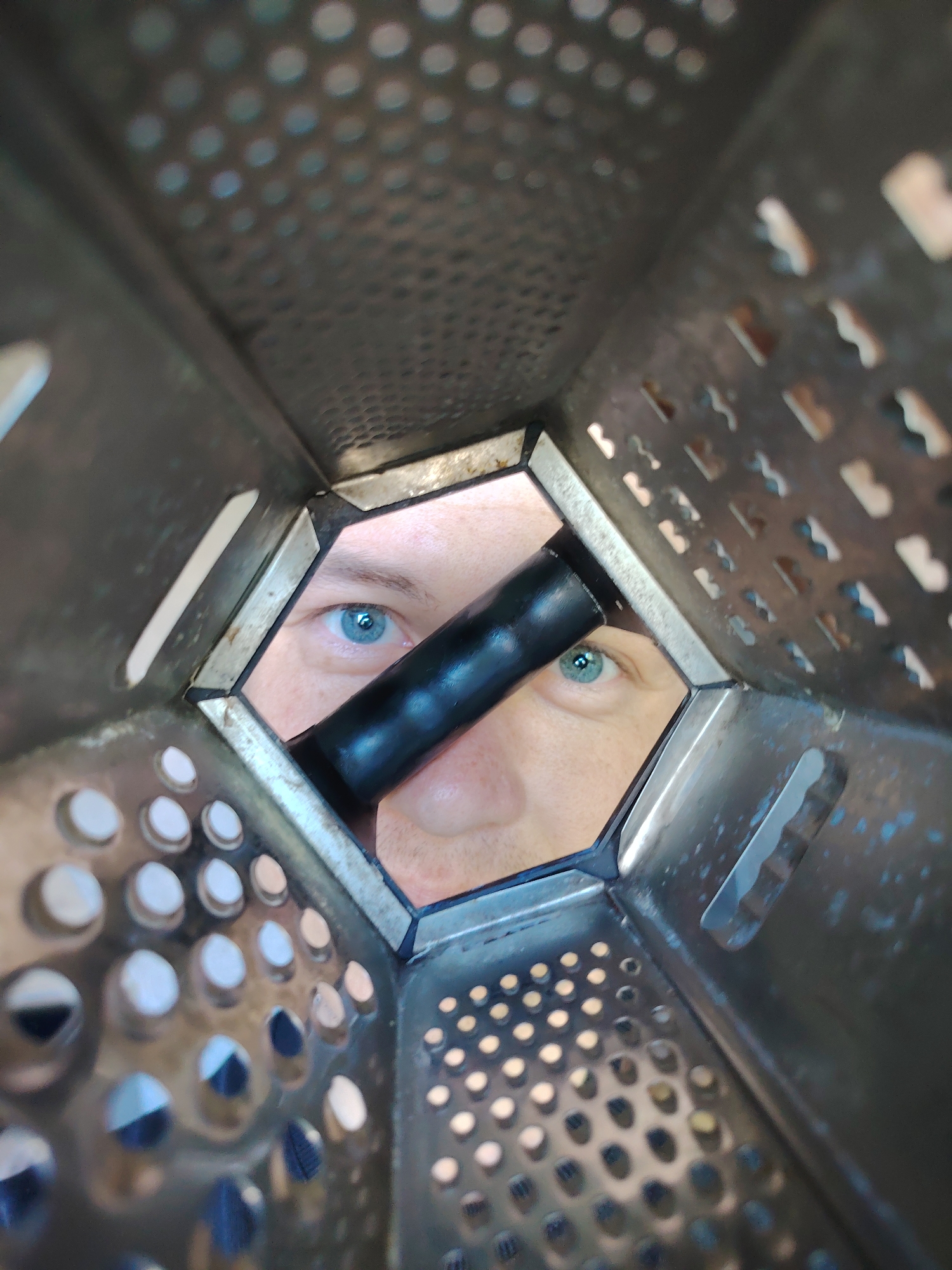The image is a close-up, extreme perspective shot looking out from the inside of a box grater. A person's face, specifically their eyes and nose, are visible through the central opening of the grater. A black handle is positioned across the opening, partially obscuring the face. The grater itself is made of metal, with various grating surfaces visible on all sides, comprised of both circular and slotted holes. The image suggests an unusual and humorous perspective, as if the viewer is peering out from within the kitchen utensil. Location and date are not identifiable from image content.