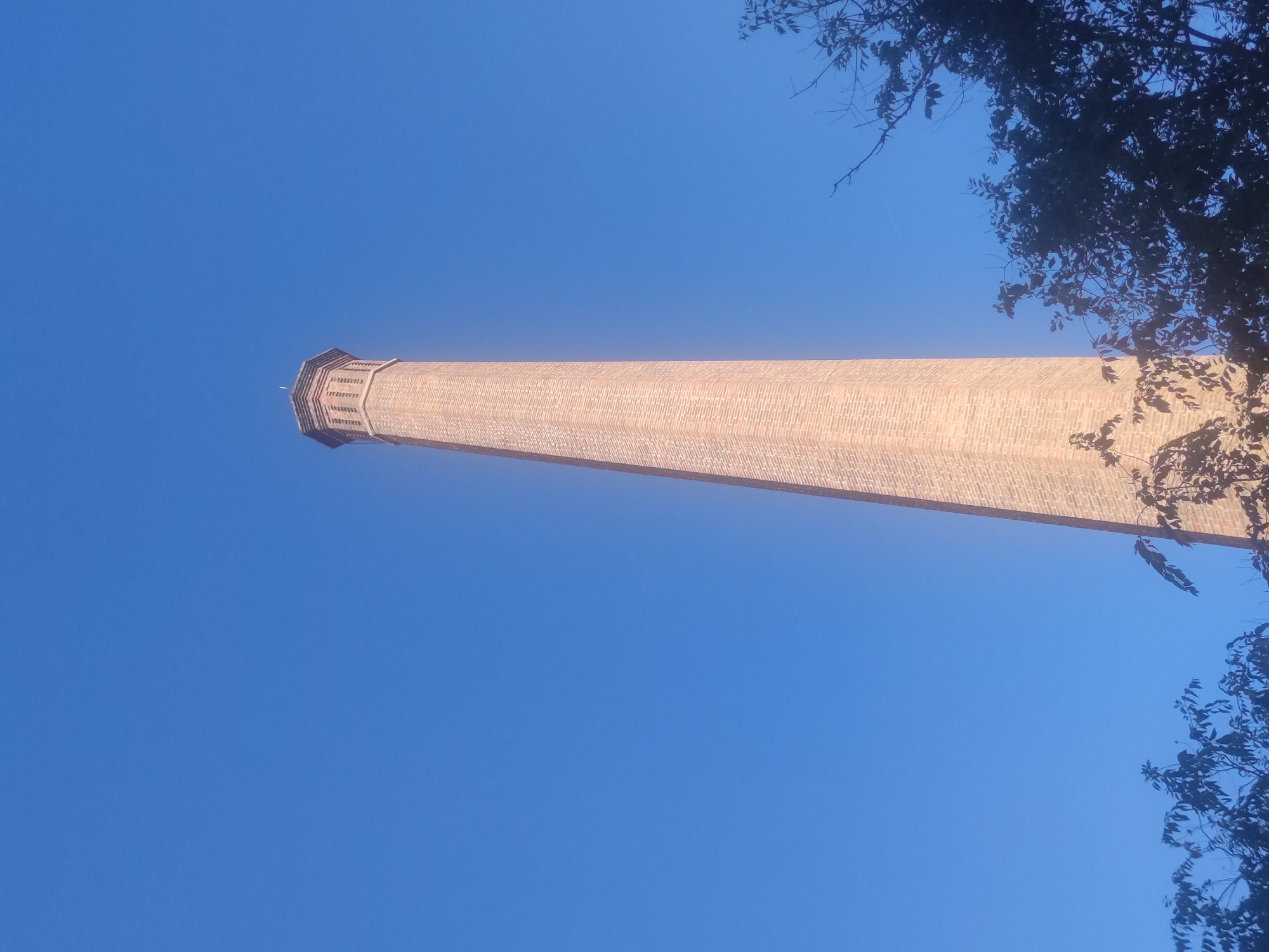 Here is a description of the image:
The image shows a tall, brick chimney or tower extending diagonally across the frame from the lower right to the upper left. The tower is composed of red bricks and has a distinct section at the top, possibly an observation deck, with decorative elements. The rest of the structure appears cylindrical and tapers slightly as it rises. The background is a clear, bright blue sky. In the upper right corner, there is a cluster of dark tree branches and leaves that partially obscure the tower. The camera angle is low, looking upward towards the tower. The location is likely the Bòvila de l'Alcalde Park in Barcelona.