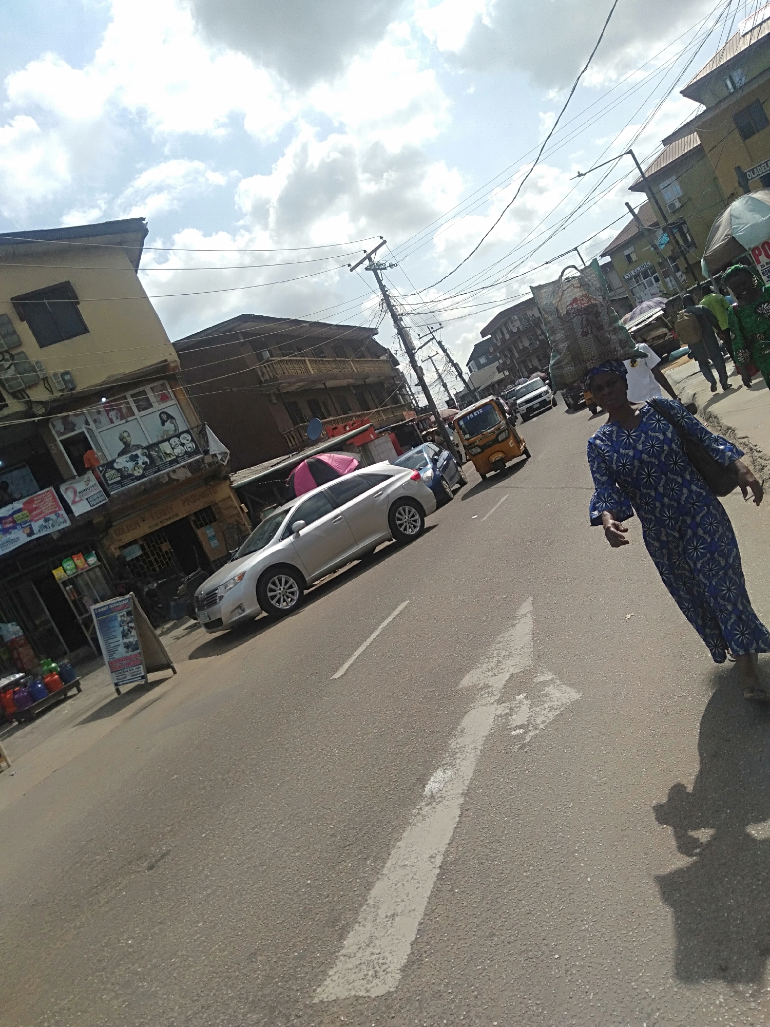 The image shows a street scene in Unknown, Unknown. A woman in a blue patterned dress walks along the street, casting a shadow. In the background, there are buildings, cars, a yellow tricycle taxi, and other people. The sky is cloudy.