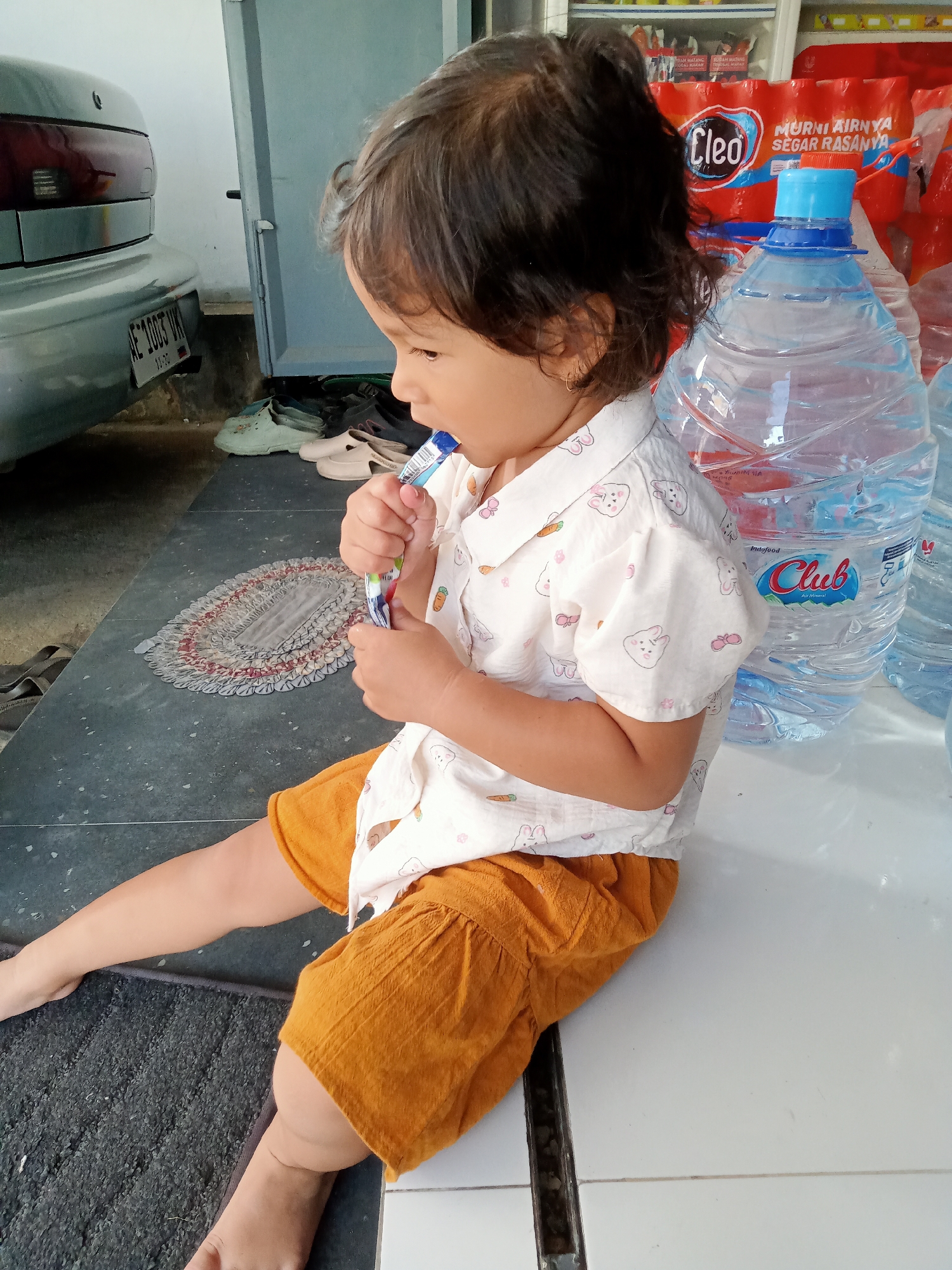 A young child, likely a girl, is seated on a white surface, possibly a table or a ledge, outdoors. She is wearing a white short-sleeved shirt with a pattern of cartoon bunnies and carrots, and orange shorts. Her left leg is extended and her right leg is bent. She is holding a small, colorful object in her hands and appears to be examining or interacting with it. Behind her, to the left, is the rear of a silver car with a license plate reading "AE 1003 VM". Further back, a light blue door is visible. To the right of the child are several large plastic bottles of water, one clearly labeled "Club". In the background, there are shelves stocked with goods, suggesting this location may be a shop or a convenience store. The ground in front of the child is a dark, tiled surface with a small, oval-shaped mat. The scene appears to be in Simo, Indonesia, as indicated by the car's license plate format which is common in the East Java region.