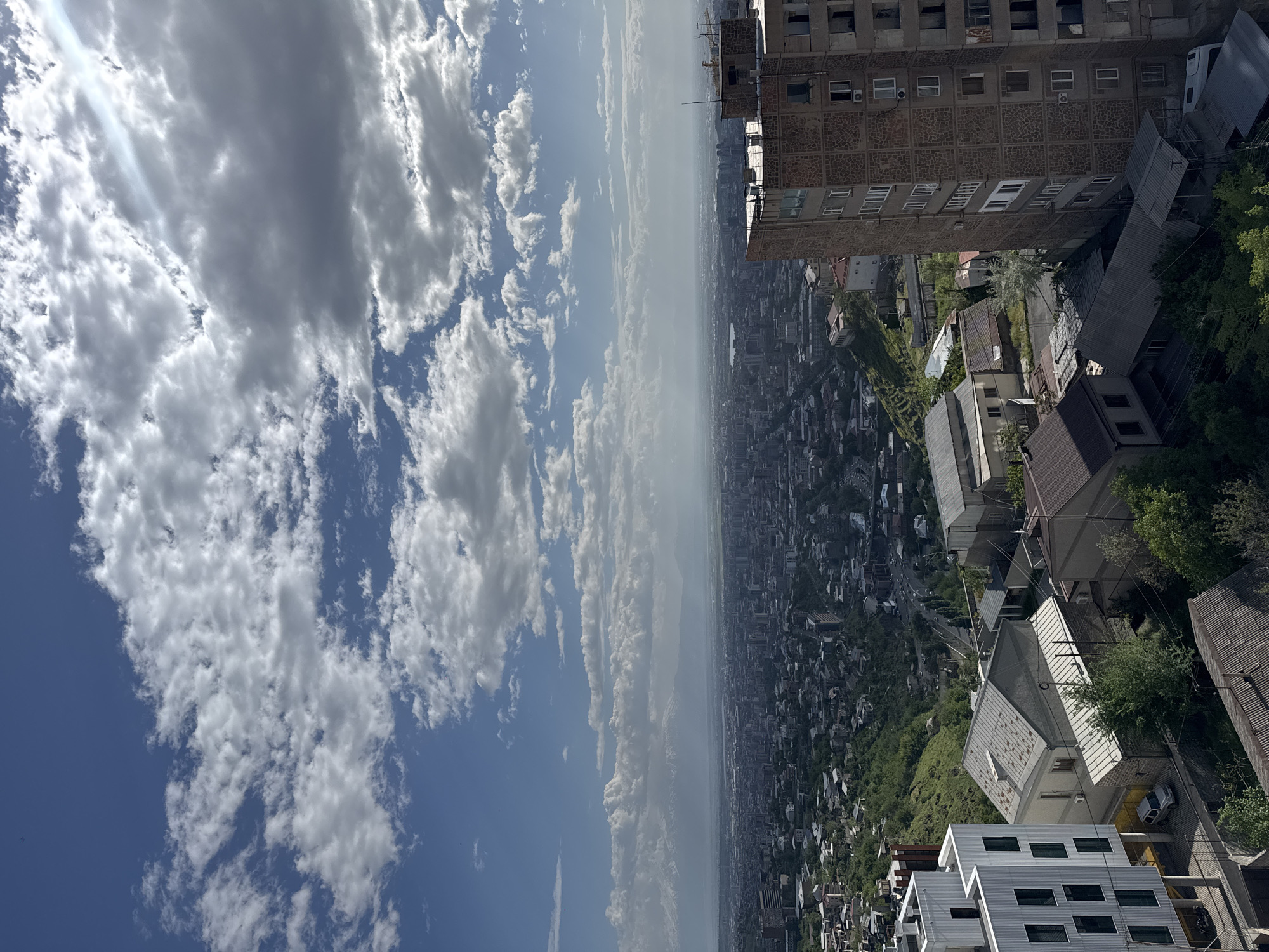 The image is a high-angle, almost aerial view of a cityscape in Yerevan, Armenia, under a partly cloudy sky.
The upper right portion of the frame shows a tall, multi-story building with a textured exterior. Below the building are various houses and smaller buildings with differing roof styles. Green foliage is visible near the buildings.
The city sprawls out in the center of the image, densely populated with buildings and structures. A haze or atmospheric distortion affects the visibility of the distant parts of the city.
The left side of the frame is dominated by a blue sky with numerous white clouds of varying density and shape. A glimpse of sunlight cuts through the clouds.