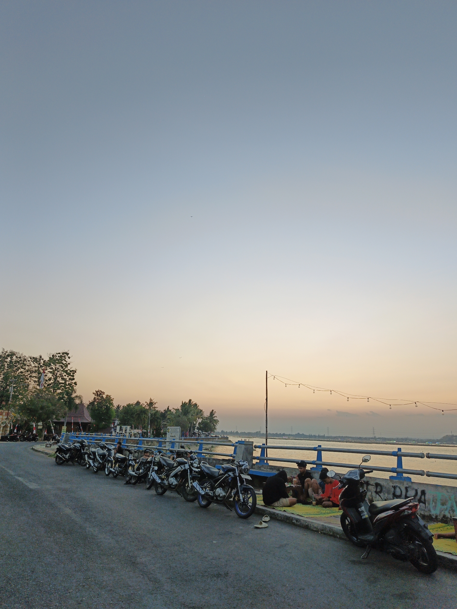 The image depicts a scene at the beachside in Krakitan, Indonesia, during a sunset. A row of parked motorcycles lines the asphalt road adjacent to a blue railing. Several people are seated on yellow mats behind the railing, facing the water. One person is wearing a red shirt, another a black hoodie, and two are in dark clothing. They appear to be socializing or relaxing. A scooter is parked in the foreground. The sky displays a gradient of blue and orange, indicating the approaching dusk. Trees and buildings are visible in the background, along with what appear to be power lines and transmission towers in the distance. String lights are suspended above the railing, adding to the ambiance of the location. The overall atmosphere suggests a casual, communal gathering at a popular seaside spot.