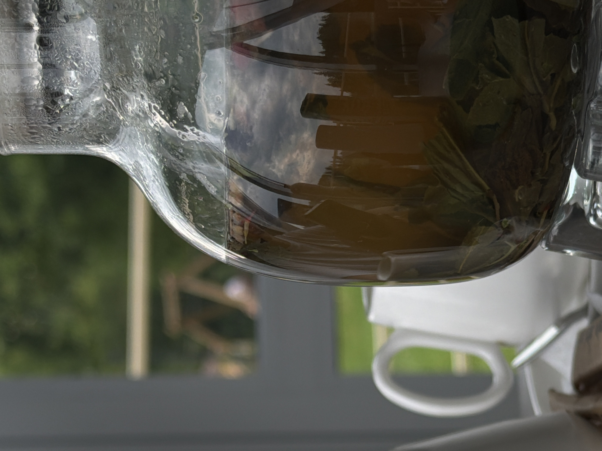 The image depicts a close-up of a glass carafe filled with a dark liquid and green leaves, likely iced tea or a similar beverage. Condensation is visible on the exterior of the carafe. To the left, a white pitcher with a curved handle is partially visible, along with a metal spoon. The background is blurred but suggests an outdoor setting with greenery and possibly a wooden structure or fence. The overall scene appears to be a relaxed, outdoor gathering. There is no indication of the city of Chusovoy, Russia.