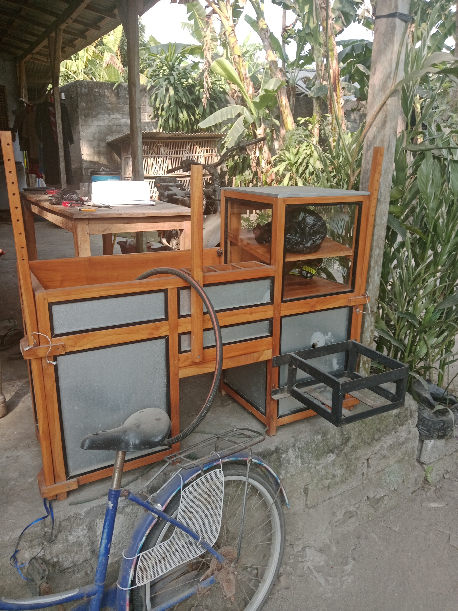 The image depicts an outdoor scene in what appears to be a residential or semi-commercial area in Sumberasri, Indonesia. The foreground is dominated by a blue bicycle with a worn, black bicycle seat. The bicycle is positioned in front of a wooden and glass display structure, possibly a stall or a cabinet. This structure has multiple shelves, some of which contain items like a black plastic bag and yellow packaging. A metal rack is attached to the lower front of the structure.
Behind the display structure, a wooden table is visible, laden with various objects including cigarette packs, a lighter, and a white laptop or notepad. A large rubber hose is draped over the wooden display unit. The background reveals a collection of tall, leafy plants, including banana trees, and a partially visible concrete wall. A corrugated metal roof and a chicken coop-like structure can also be observed, suggesting a rural or semi-rural setting. The overall impression is one of an unkempt, functional outdoor space.
