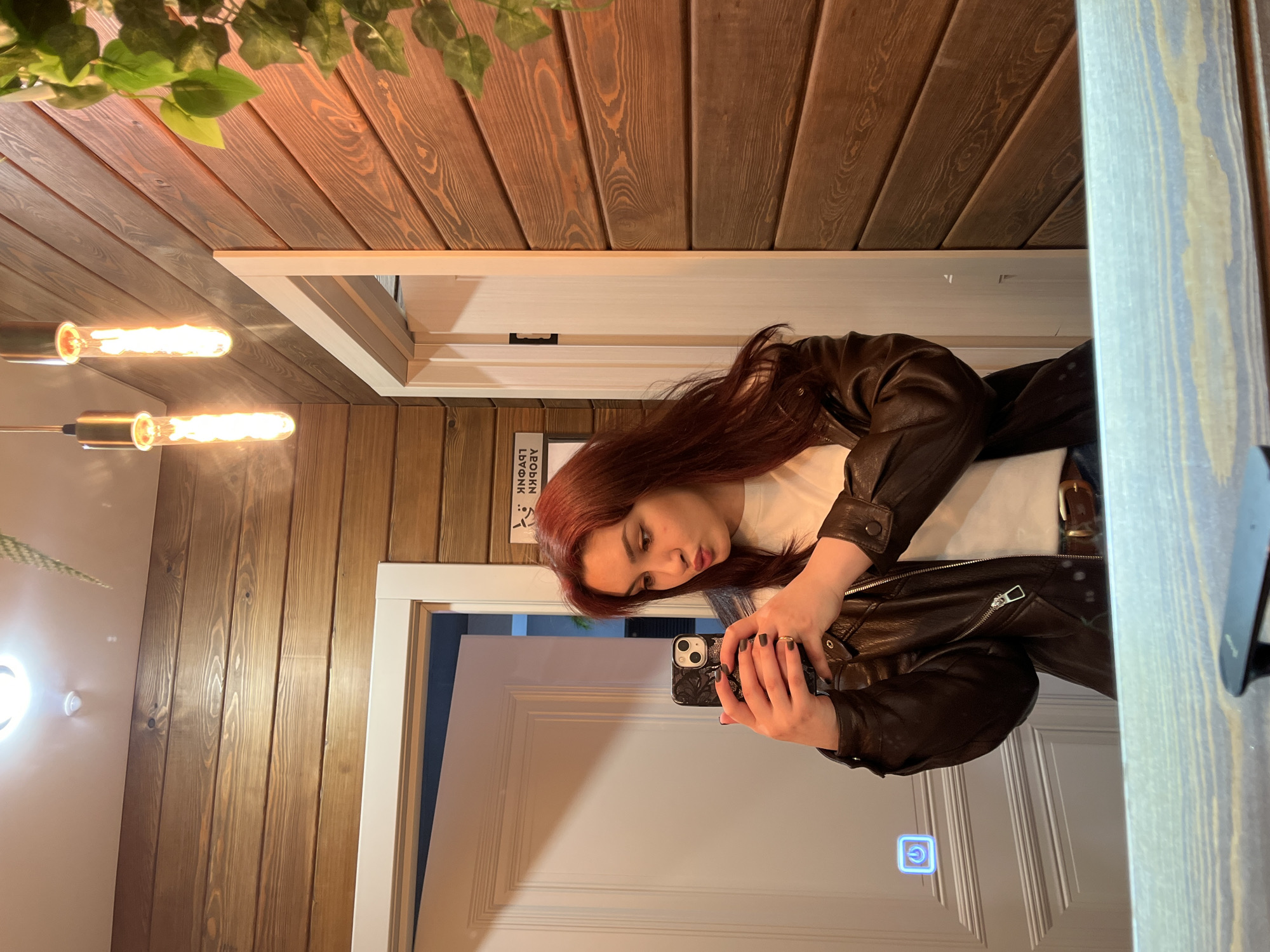 The image depicts a young woman in front of a mirror, presumably taking a selfie with her phone. She has red hair and is wearing a white t-shirt and a dark leather jacket. The reflection shows a white door with a blue power button on it. The setting includes walls with brown wooden paneling and hanging plants. A sign with Cyrillic text is visible on the wall near the door frame. Additional light fixtures with elongated bulbs are also visible. While it is challenging to pinpoint the exact location within the city of Neftekamsk, Russia, based on this image alone, the wooden paneling suggests an interior setting, potentially a cafe, bar, or other establishment.