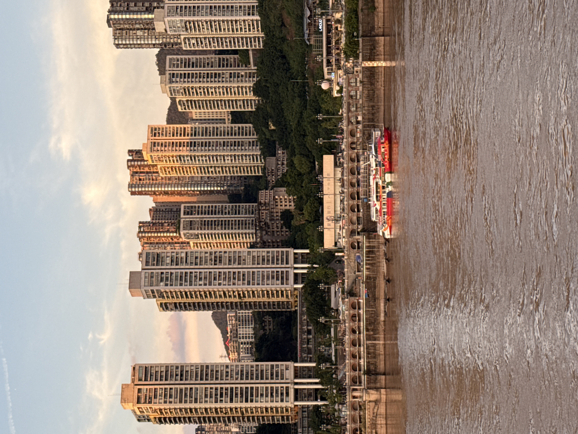 The image depicts a cityscape along a river, likely in Yuzhong District, China, during what appears to be late afternoon or early evening, given the warm lighting. The foreground is dominated by a muddy, rippling river. In the middle ground, a multi-level embankment lines the river. The lower level of the embankment is a stone wall with arched openings. Above this, a wider promenade or road is visible, with trees and lampposts lining it. Several modern, high-rise buildings rise steeply from behind the embankment and the lush green hillside, dominating the background. These buildings are tall and slender, with many windows. A red and white passenger boat is docked at the embankment, its reflection visible in the water. The sky above is a mix of blue and pale orange, with scattered wispy clouds, suggesting a sunset. The overall impression is one of a densely populated urban environment situated along a significant waterway, with development cascading up the hillsides.