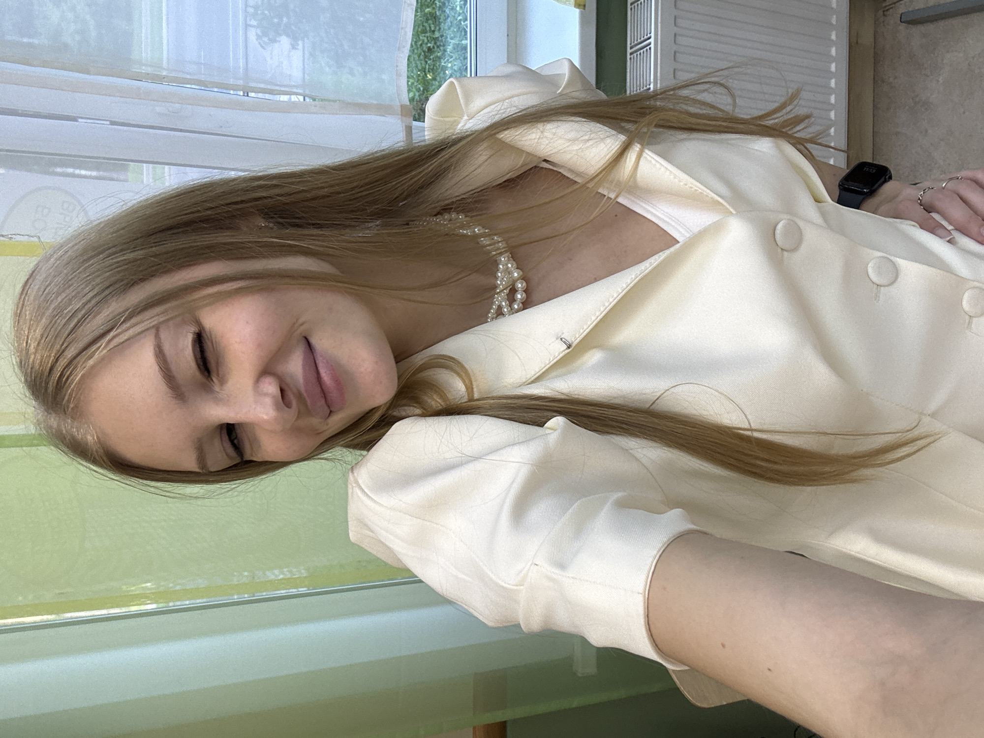 A young woman with long, blonde hair is taking a selfie. She is wearing a cream-colored blazer over a white top and a pearl necklace. Her right eye is closed in a wink, and she is smiling. The background shows a green curtain and a window with a view of trees and a street. A white radiator is visible below the window. She is wearing a smartwatch on her left wrist. The scene appears to be indoors, possibly in an office or a room with a view. There is no discernible information that would suggest this location is in the city of Perm, Russia.