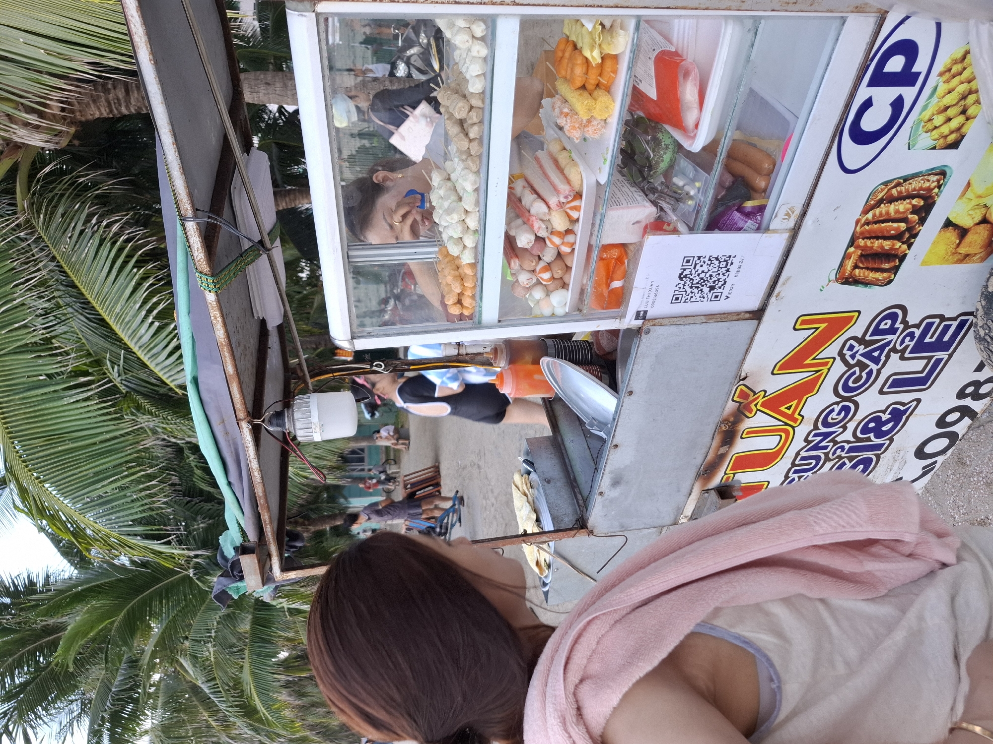 The image shows a street food stall in Bac Nha Trang Ward, Vietnam. The stall is a glass-fronted refrigerated display filled with various skewers of food, including meatballs, sausages, and seafood. A woman, likely the vendor, is visible behind the stall, looking down at her food. In the foreground, a person is seen from the back, wearing a pink towel draped over their shoulder. The background suggests a beachside location with palm trees and other people in casual attire. The presence of palm trees and the general atmosphere indicate a warm, tropical environment, likely daytime. A QR code is visible on the side of the stall, along with text that appears to be in Vietnamese. The overall impression is of a casual, everyday scene at a popular tourist spot.