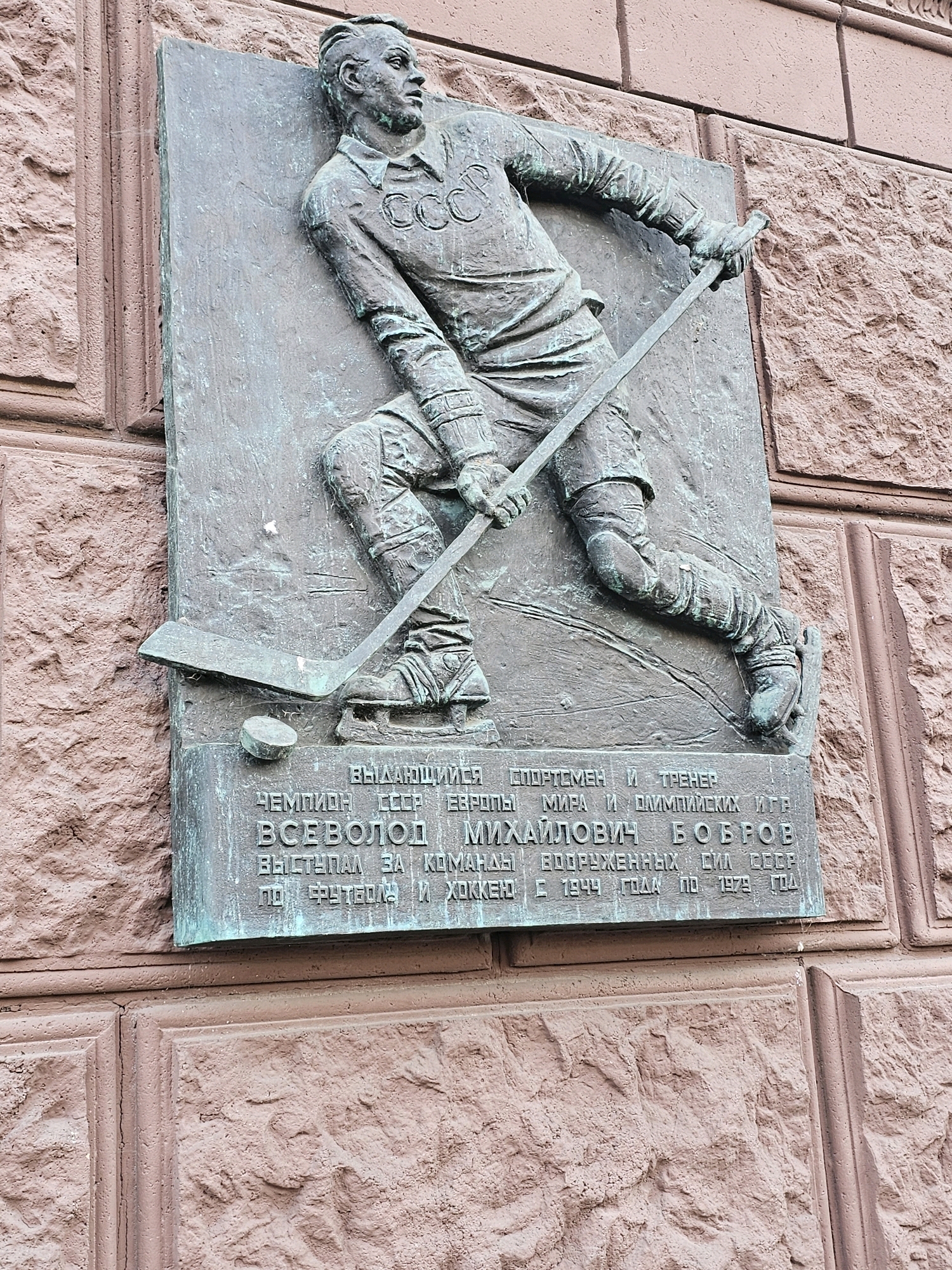This is a close-up, eye-level, outdoor, medium shot of a bronze relief plaque mounted on a textured reddish-brown brick wall. The plaque depicts a male hockey player in mid-action, with his body leaning forward as if skating. He is wearing a hockey uniform with "CCCP" emblazoned across the chest and a helmet. He is holding a hockey stick, which is positioned near a puck on the ice. Below the hockey player, there is an inscription in Cyrillic text. The plaque is tarnished and has a greenish patina. The brick wall is rough and irregular in texture. The lighting suggests it is daytime, possibly overcast, casting soft shadows. The location is identified as being in Khimki, Russia.