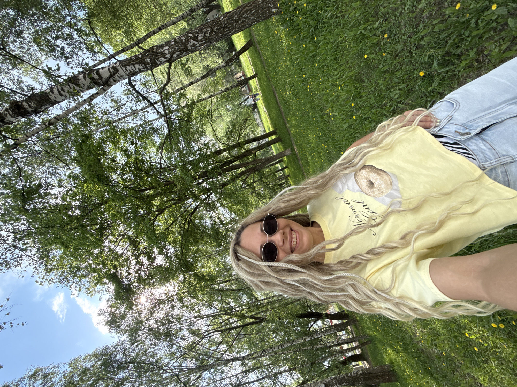 A woman is lying in a grassy area in Rusanovo, Russia. She is wearing sunglasses, a yellow shirt with a donut on it, and light blue jeans. Her hair is blonde and styled with white braids. The grassy area is dotted with yellow flowers and surrounded by trees. The sky is visible through the trees.
