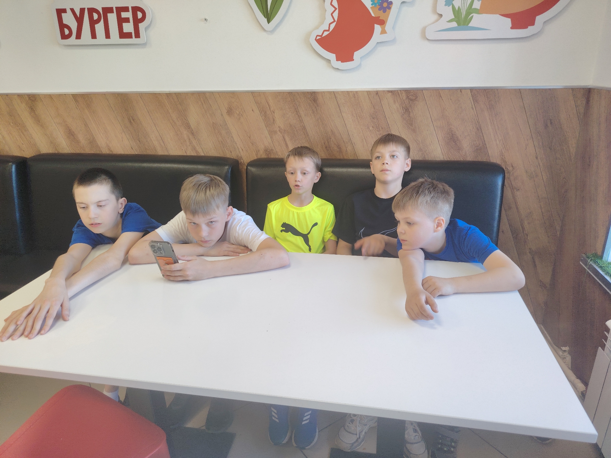 Five boys are seated at a white table against a black upholstered booth in what appears to be a casual dining environment. The boy on the left is wearing a blue shirt and has his arms resting on the table. Next to him, a boy in a white shirt is looking at a phone. The middle boy is wearing a bright yellow Puma shirt. To his right is a boy in a black shirt looking upwards and outwards. The boy on the right is wearing a blue shirt and resting his chin on his hand, looking to the side. Under the table, shoes are visible. Above the boys is a wall with wood paneling and, above that, a white wall with cartoonish food decorations and the word "БУРГЕР" (BURGER) in red Cyrillic letters. The location is in the city of Kungur, Russia.