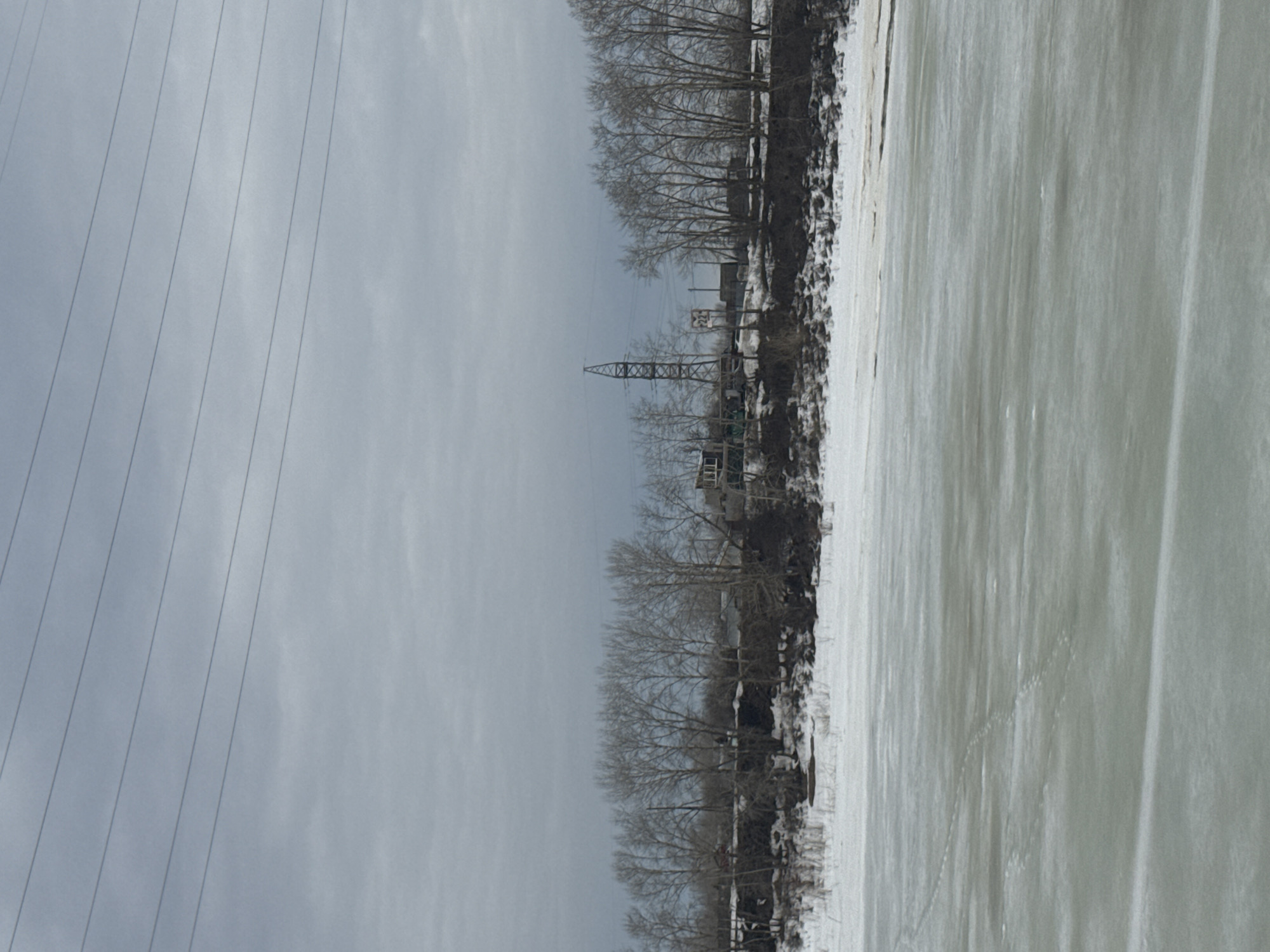 The image depicts a winter scene in what is likely Chaikovsky, Russia, based on the presence of an industrial structure along the river. An expanse of frozen water, presumably the Kama River, covers a large portion of the frame, showing traces of footprints on its surface. Along the riverbank, there are bare trees and evidence of snow cover on the ground. An electricity pylon is visible along the river bank. To the left of the river, there are utility lines in the sky. The sky is overcast, casting a diffuse light over the scene. The overall impression is that of a cold, desolate, industrial landscape in winter.